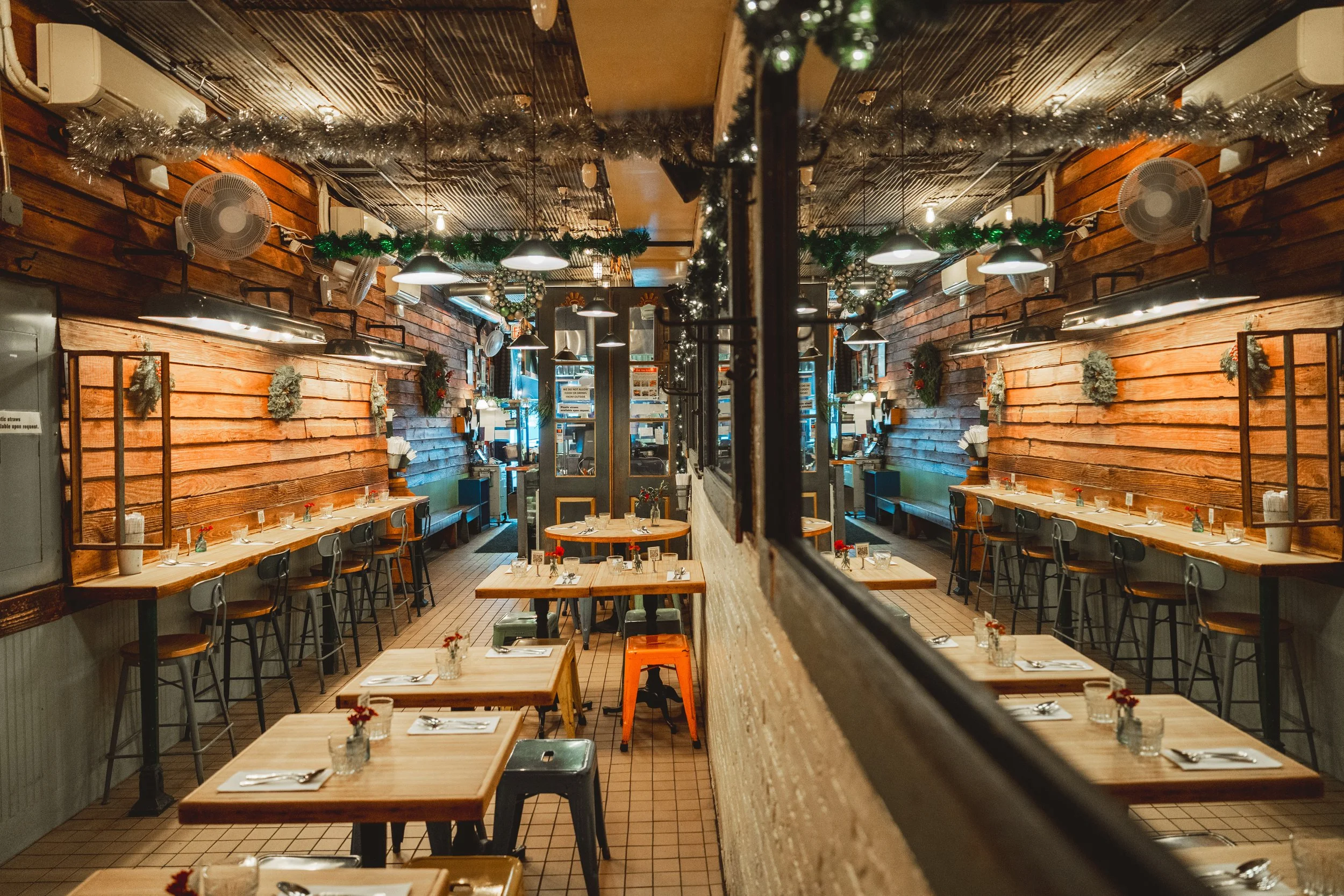 Dining room with warm wooden plank walls, bar-height counter seating, light wood tables, and festive garland strung along the ceiling.