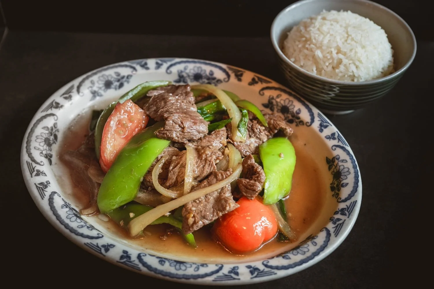 Stir-fried beef with green pepper, tomato, and onion in a savory brown sauce on a blue and white floral plate, served with a bowl of steamed rice.