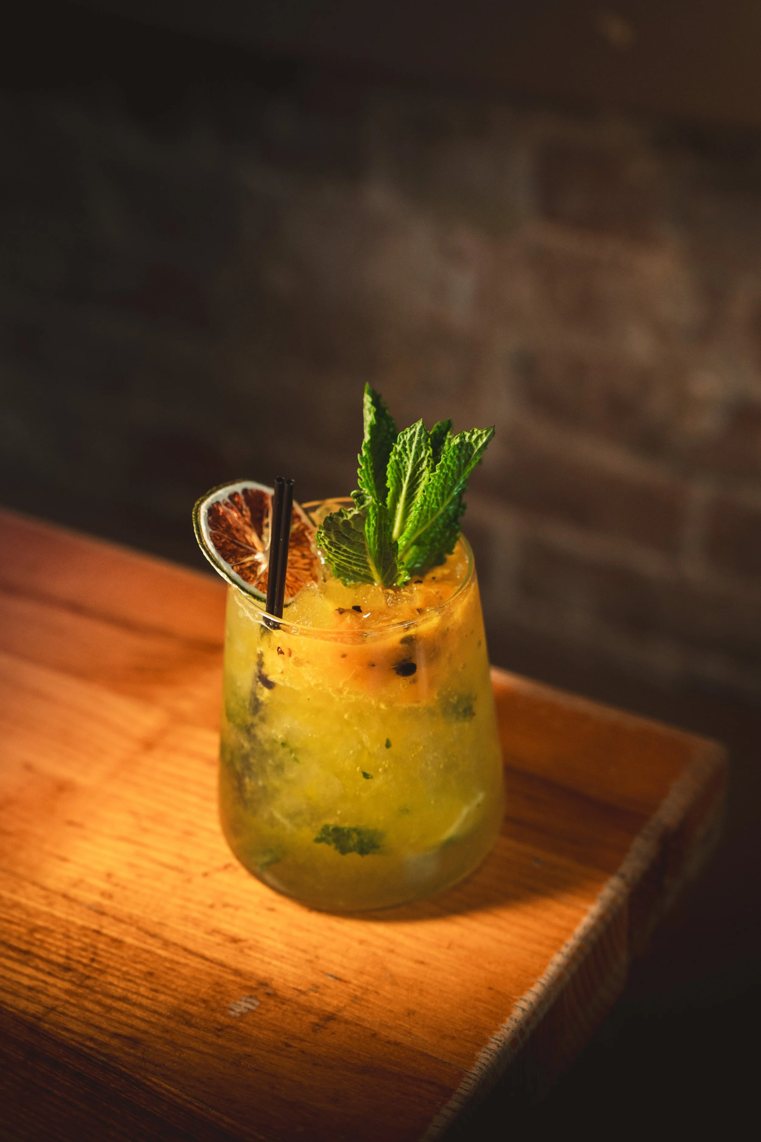 Close-up of a yellow fruit cocktail over crushed ice, garnished with fresh mint sprigs and a dried citrus slice, served with a black straw on a wooden surface.