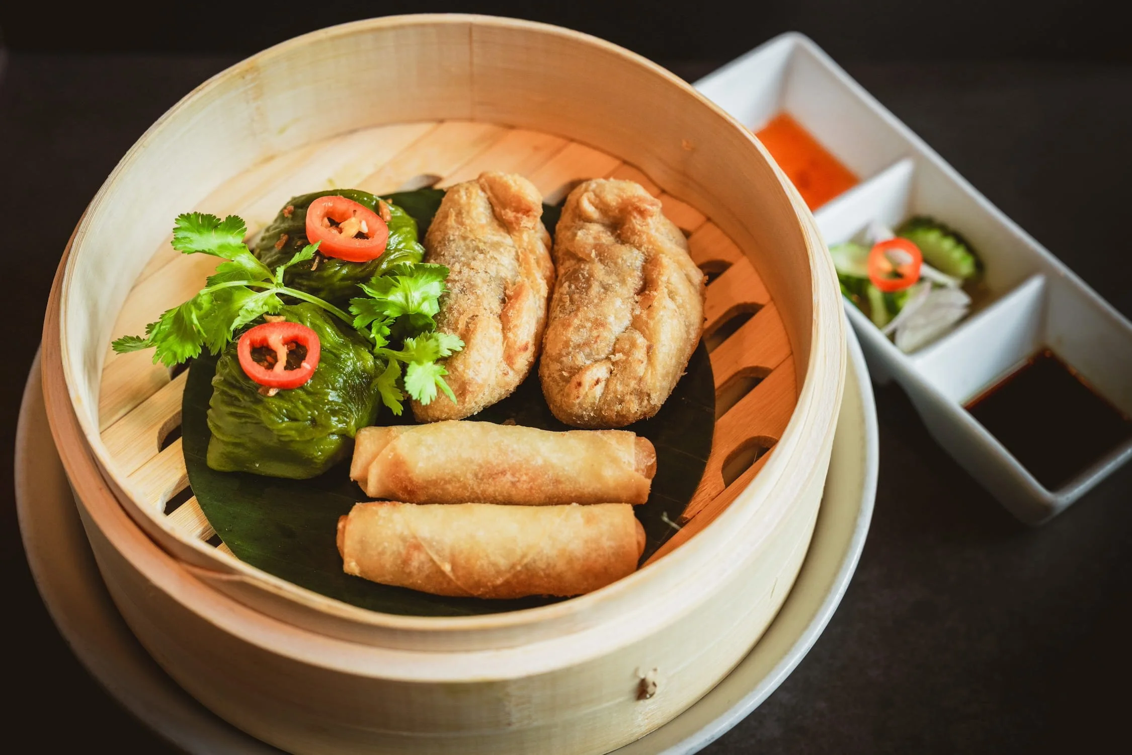Mixed Appetizers in a bamboo steamer with 2 crispy root spring rolls, 2 vegetable spring rolls and 2 green dumplings garnished with cilantro and sliced red chili, served with dipping sauces on the side.