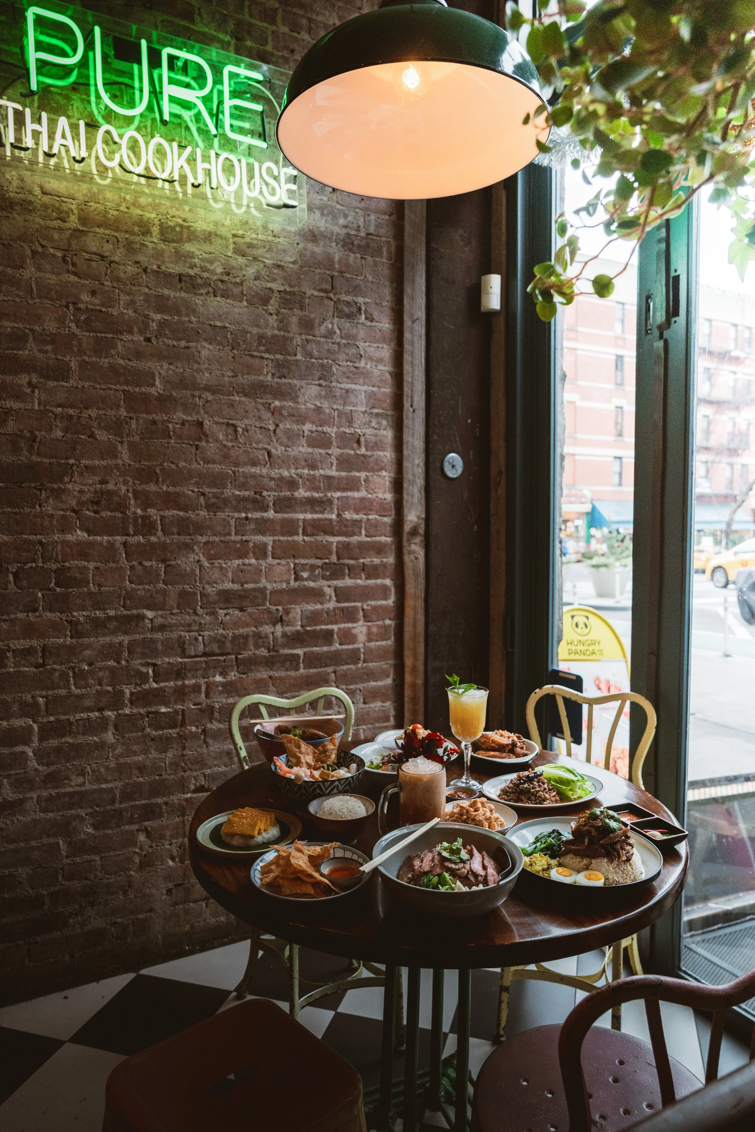 Corner table at Pure Thai Cookhouse loaded with shared dishes and cocktails, beside a large window overlooking the street, with exposed brick walls and a green neon sign above.