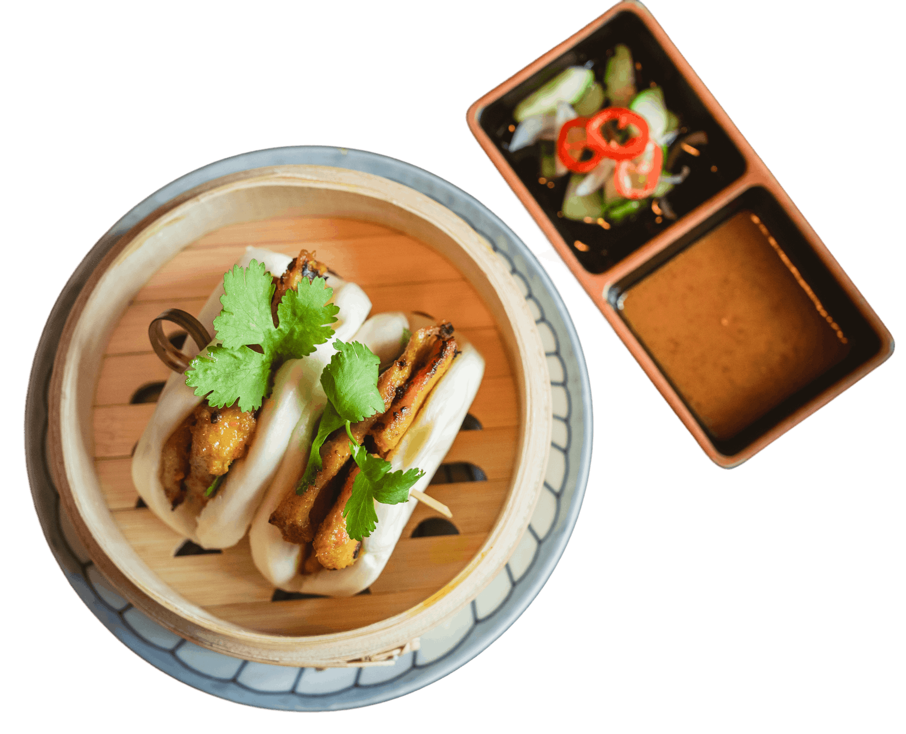 Bottom left is two chicken satay buns in a bamboo steamer and garnished with cilantro. Top right is peanut sauce and cucumber relish.