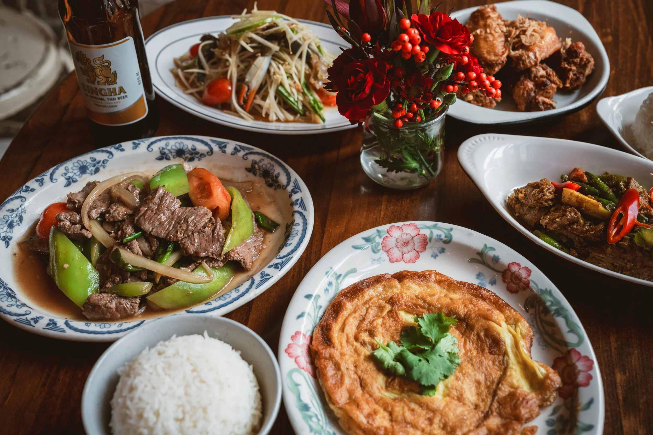 Thai family-style meal with beef stir-fry, omelette, papaya salad, rice, and Singha beer.