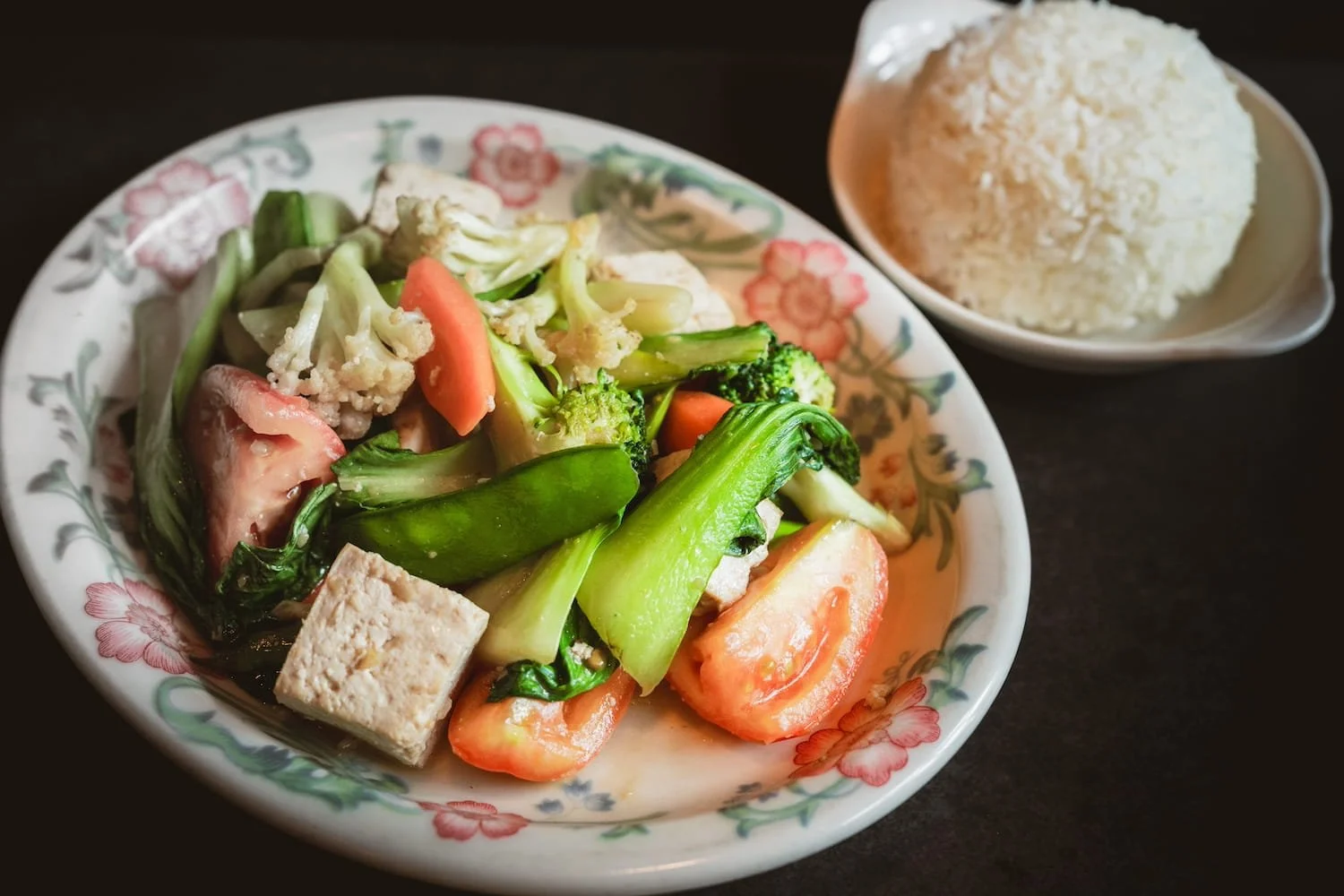 Stir-fried mixed vegetables with tofu, broccoli, bok choy, cauliflower, and tomato on a floral ceramic plate, served with steamed rice.