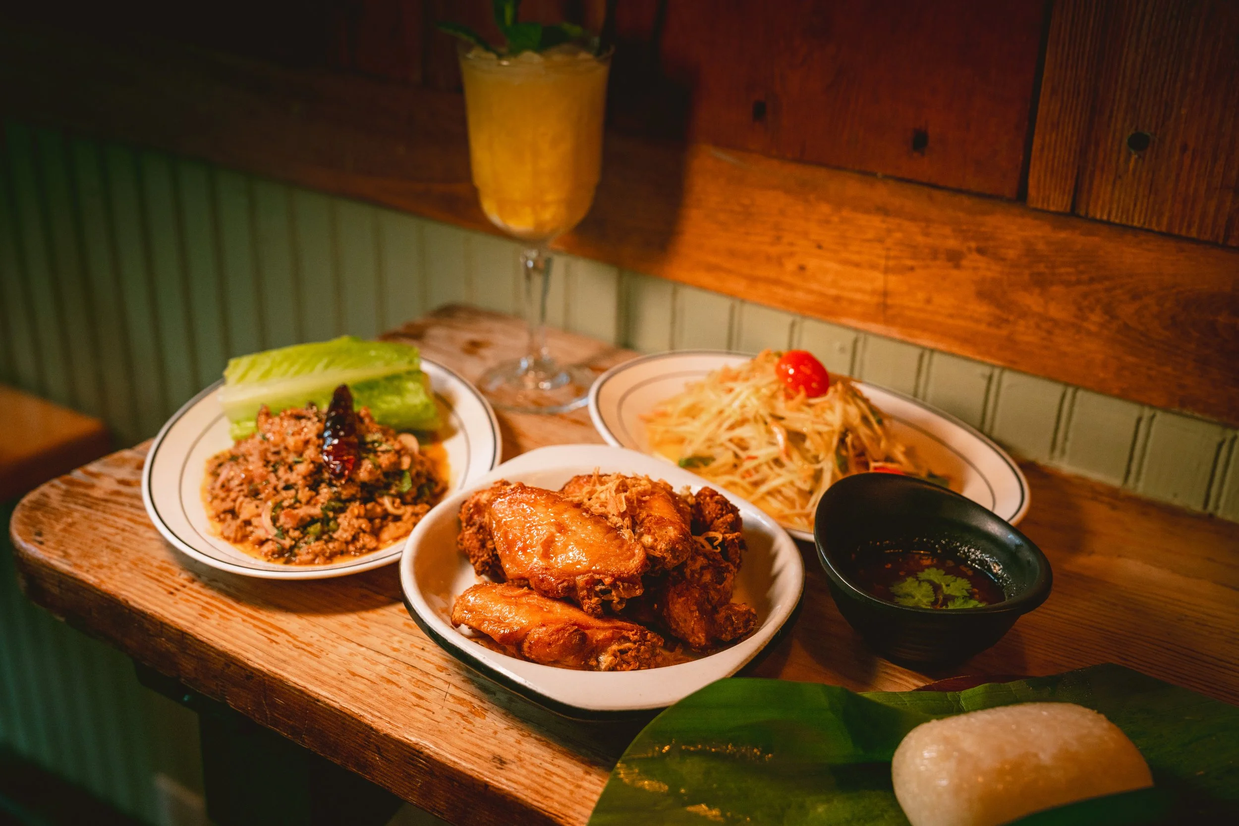 Spread of Thai dishes on a wooden table including fried chicken wings, minced meat larb with lettuce, green papaya salad, and a dark dipping sauce, with a glass of yellow drink in the background.