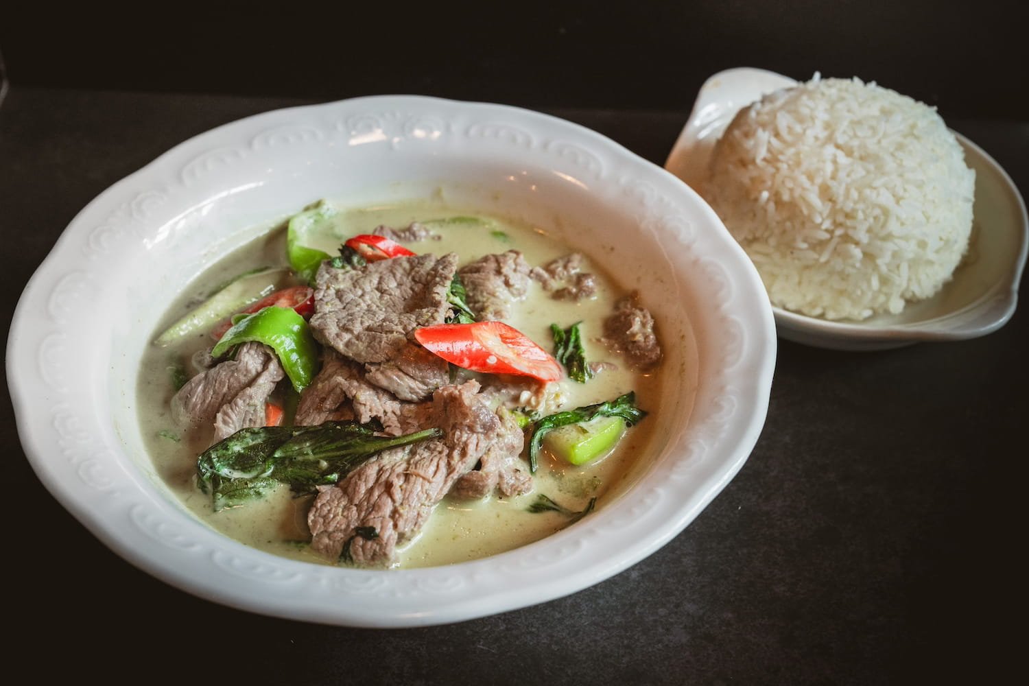 Green Curry with sliced beef, Thai eggplant, asparagus, and red chili in a creamy coconut broth, served in a white bowl with steamed rice.