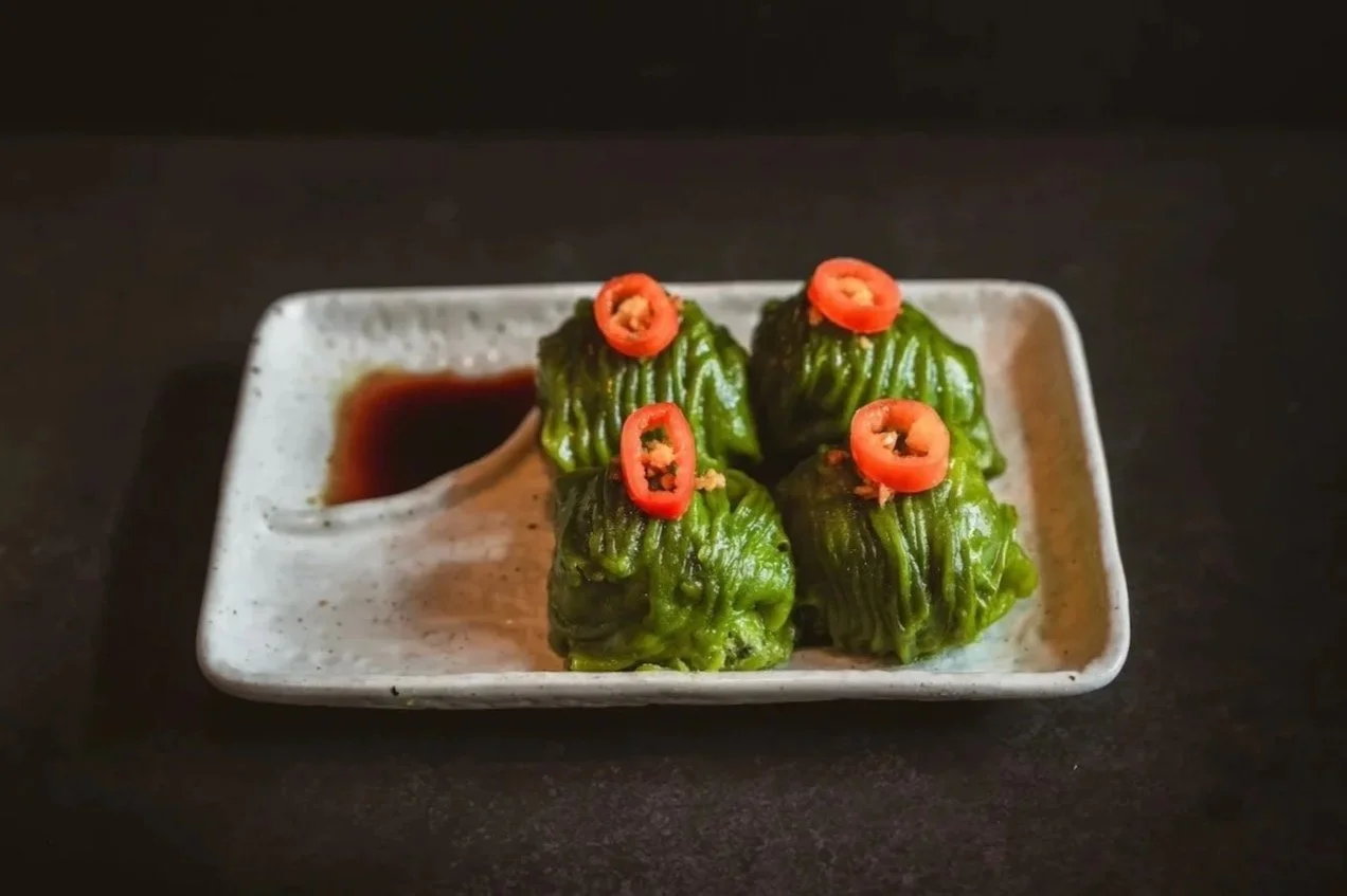 Four green steamed dumplings topped with sliced red chili, served on a rectangular ceramic plate with a dark dipping sauce.