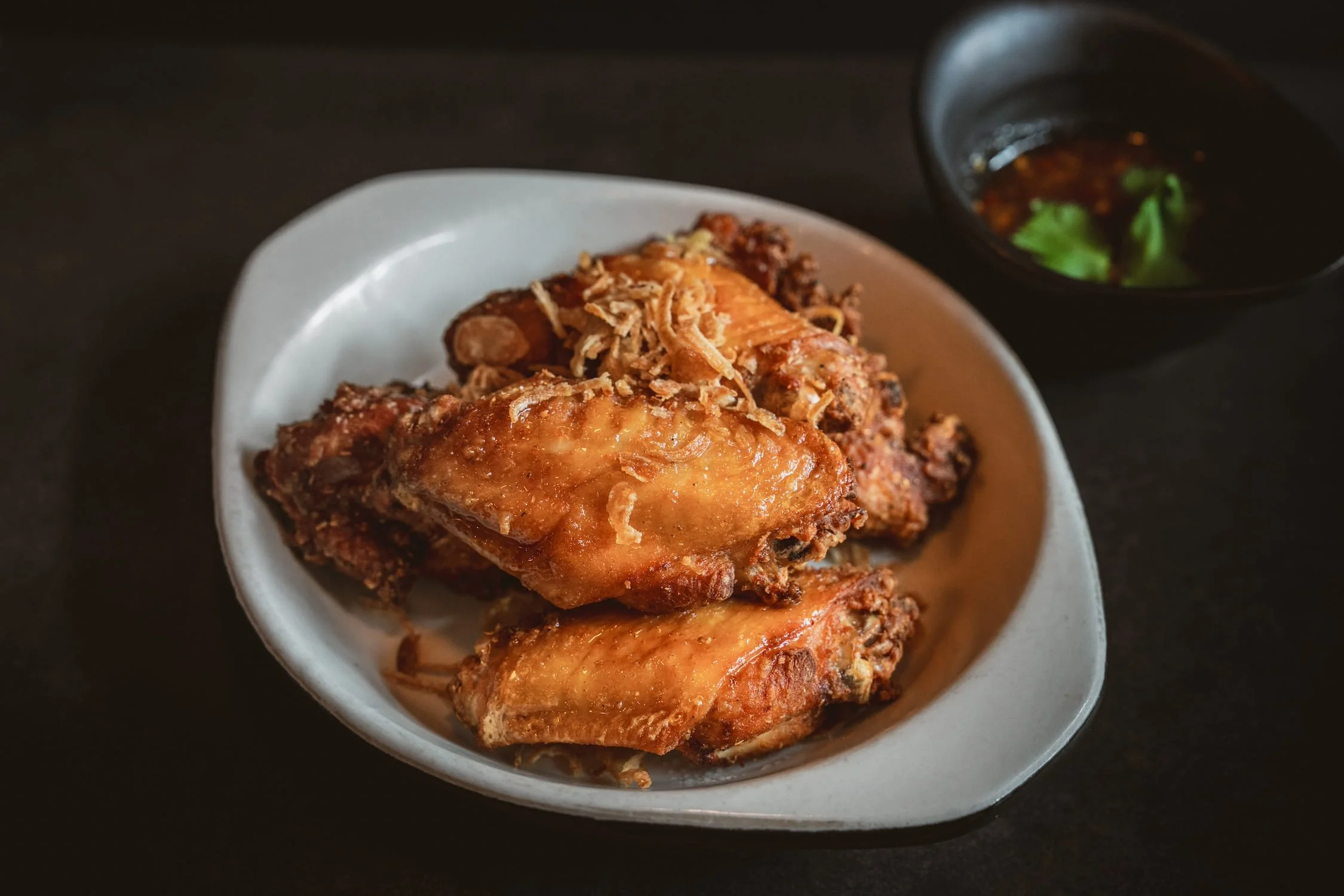 Crispy fried chicken wings topped with fried shallots in a white oval dish, with a dark dipping sauce in the corner.