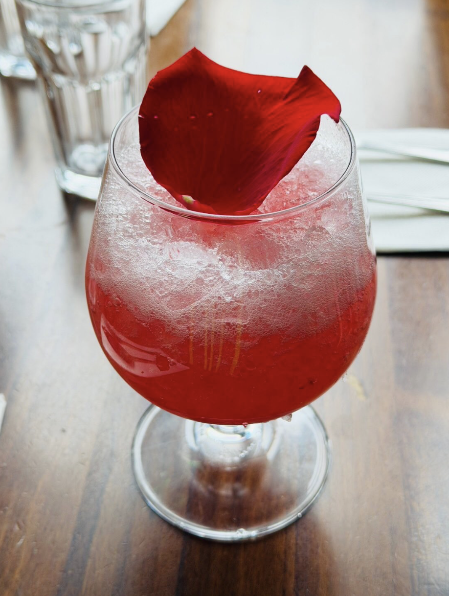 Sparkling ruby-red drink in a large wine glass, garnished with a fresh red rose petal.