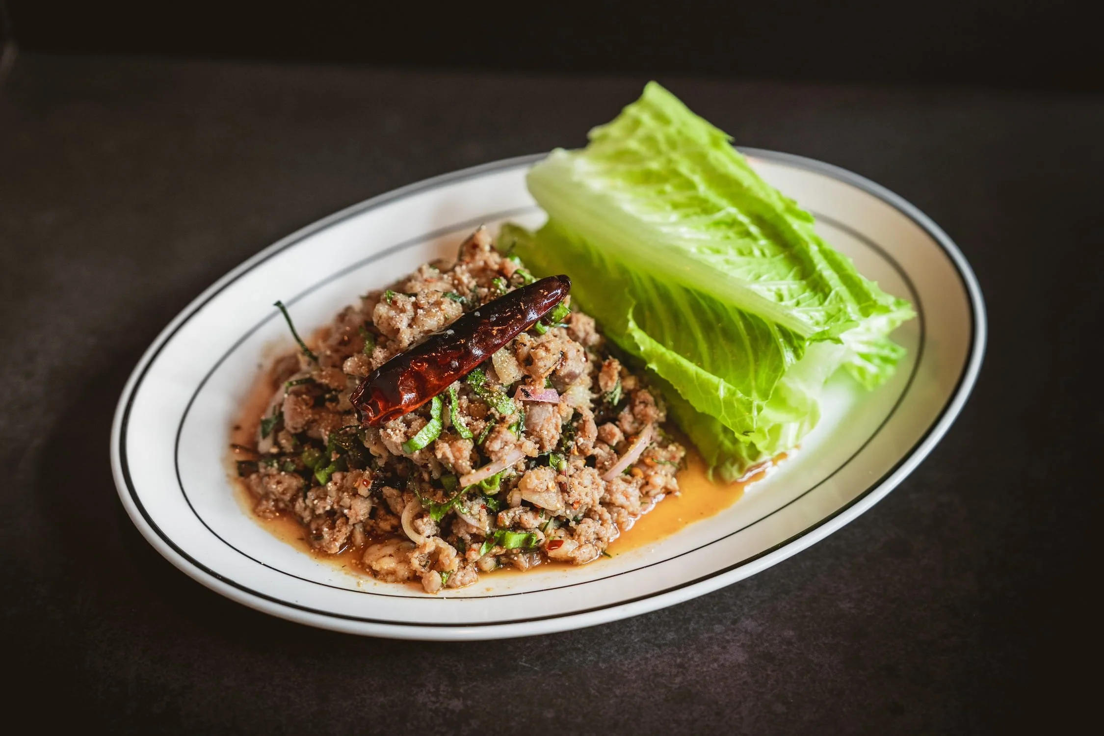 Larb Duck with minced duck, dried chili, and fresh herbs served alongside crisp lettuce leaves on an oval white plate.