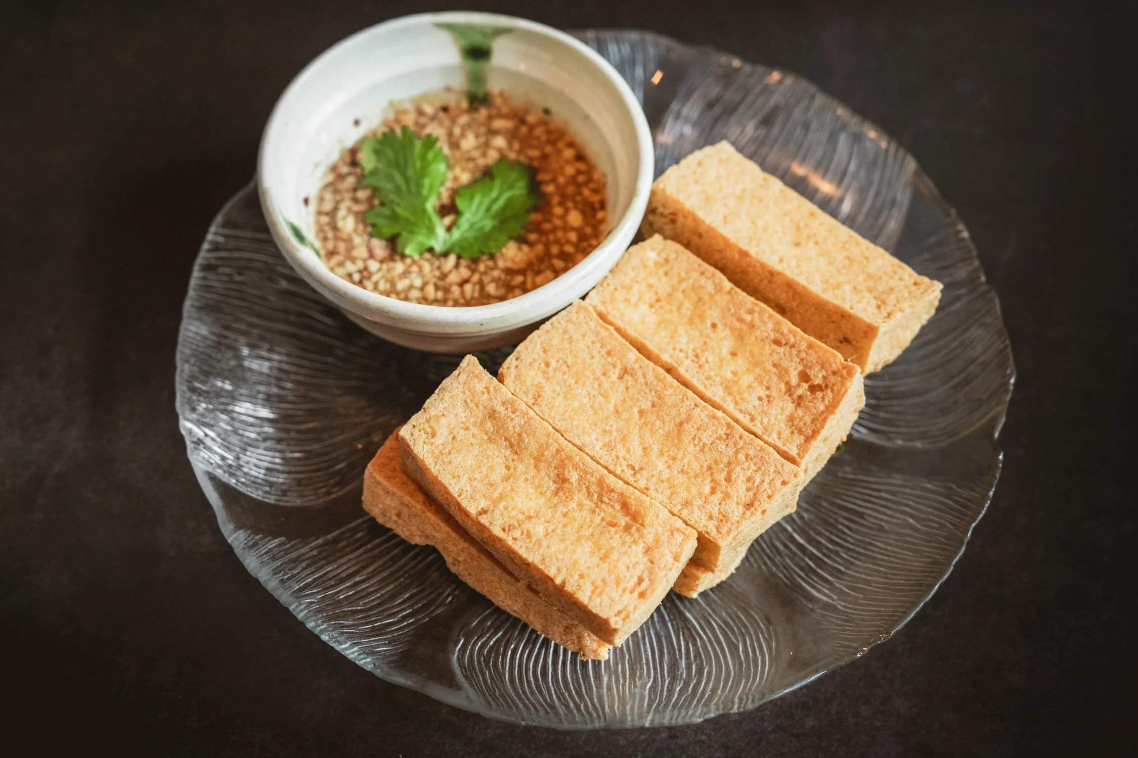Four golden-fried tofu slices on a dark plate, served with a sesame dipping sauce garnished with cilantro.
