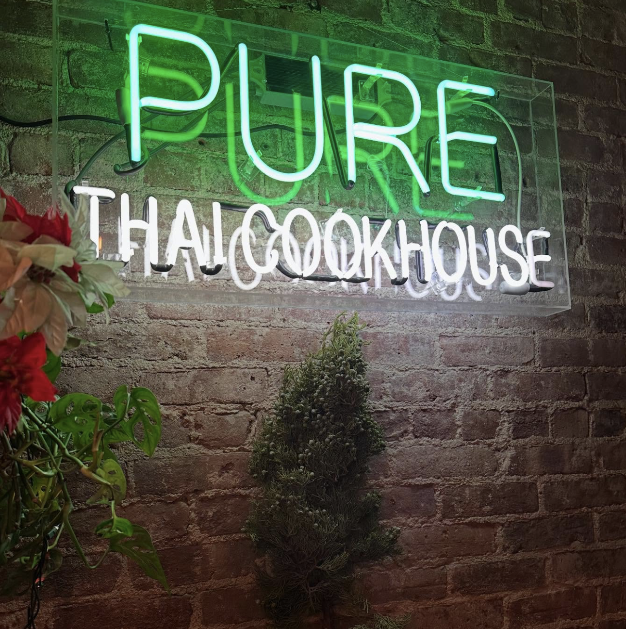 Pure Thai Cookhouse neon sign mounted on an exposed brick wall, with green lettering for PURE and white lettering for Thai Cookhouse, flanked by small plants and red flowers.