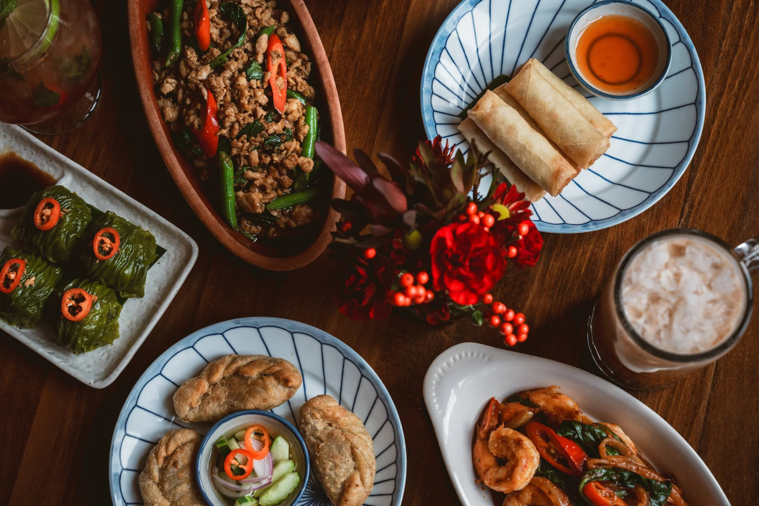 Thai dishes spread on a wooden table including crispy spring rolls, root vegetable puffs, steamed vegetable dumplings and wok basil.