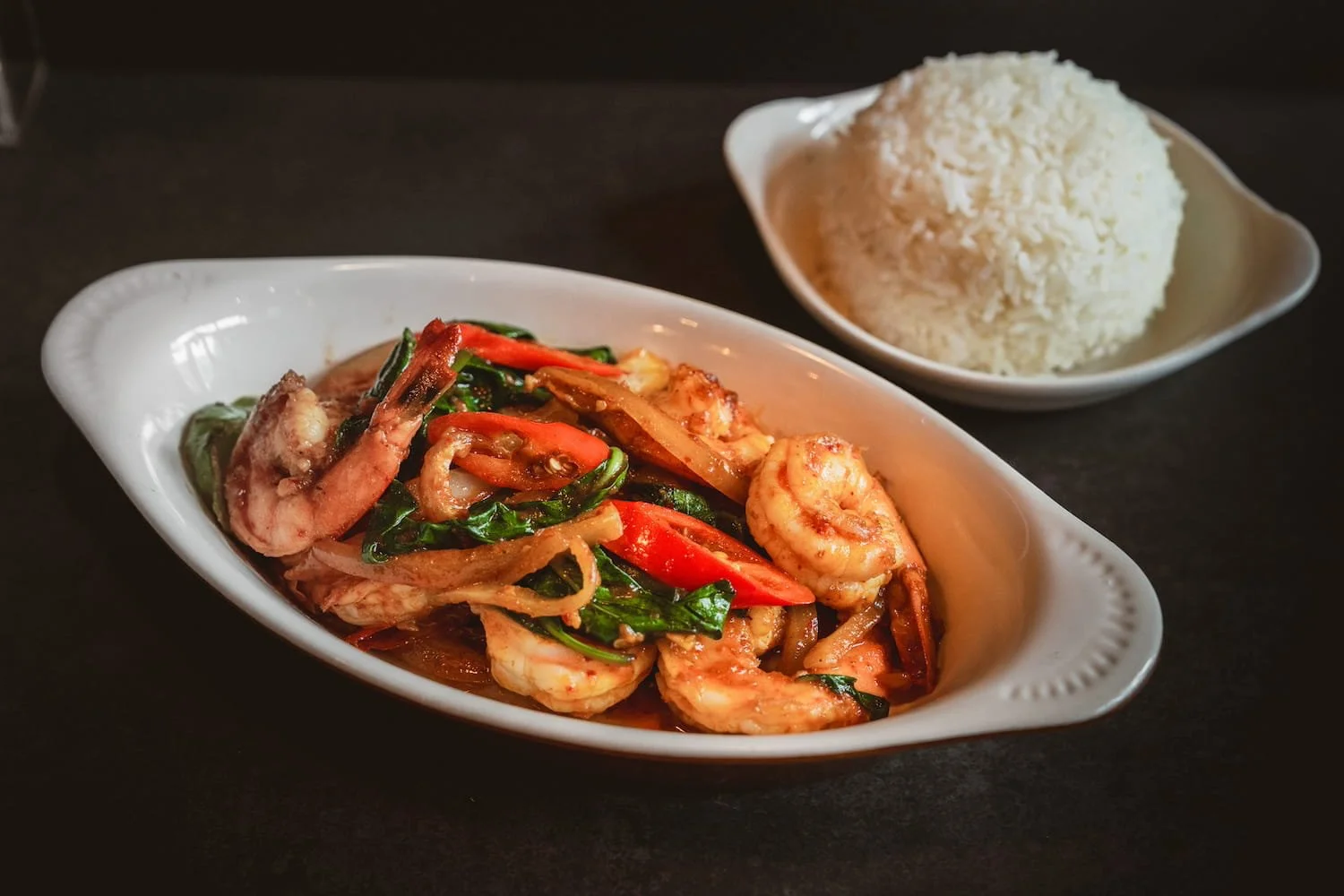 Wok Roasted Chili with stir-fried shrimp, Thai basil, onion, and long red chili in a white oval dish, served with steamed rice.