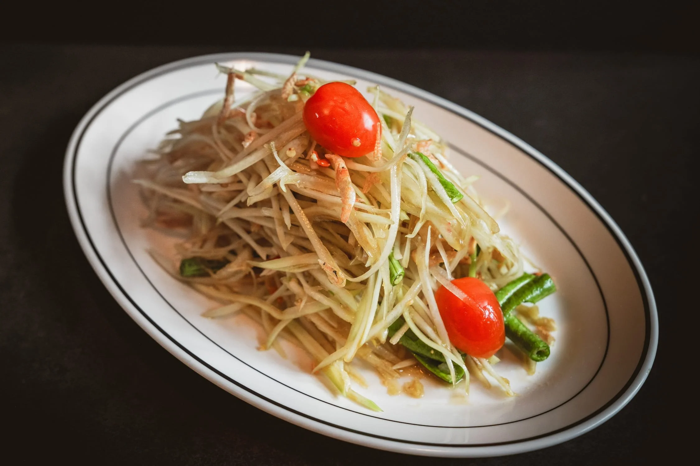 Papaya Salad with shredded green papaya, cherry tomatoes, and long beans on an oval white plate.