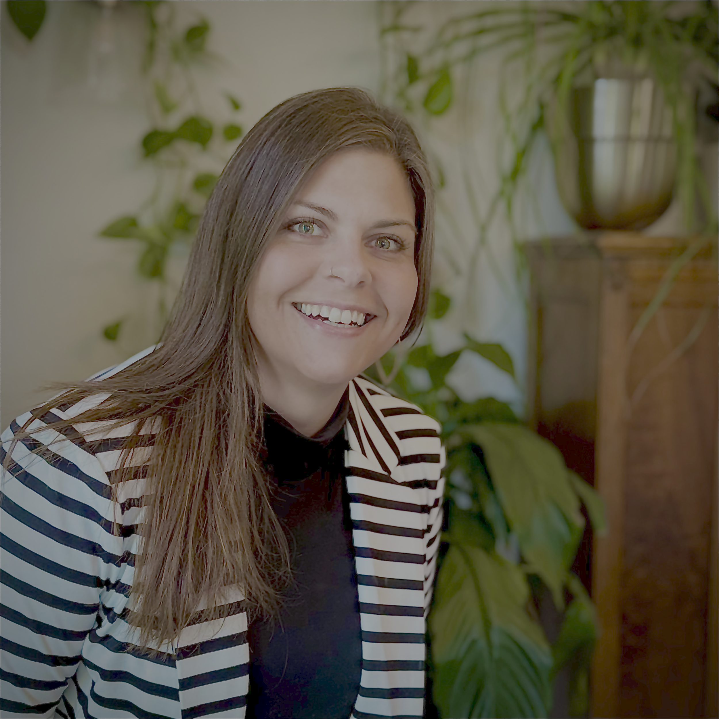 A woman smiling, wearing a black top and a black and white striped blazer, seated indoors with green plants and wooden furniture in the background.