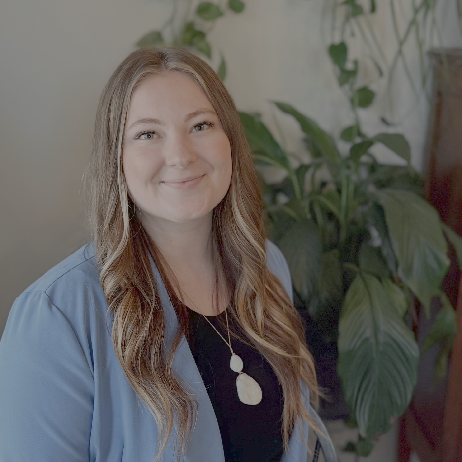 A smiling woman with long, wavy light brown hair, wearing a black top, a light blue blazer, and a statement necklace, standing in front of a large green houseplant.