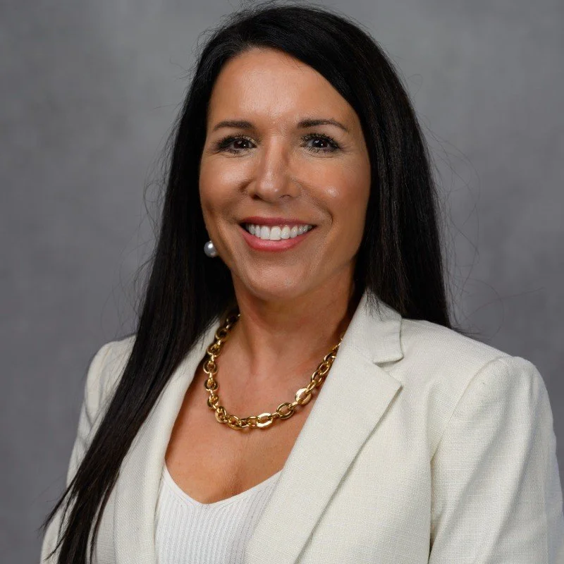 Professional woman with dark hair smiling, wearing a white blazer, pearl earring, and gold chain necklace against a gray background.