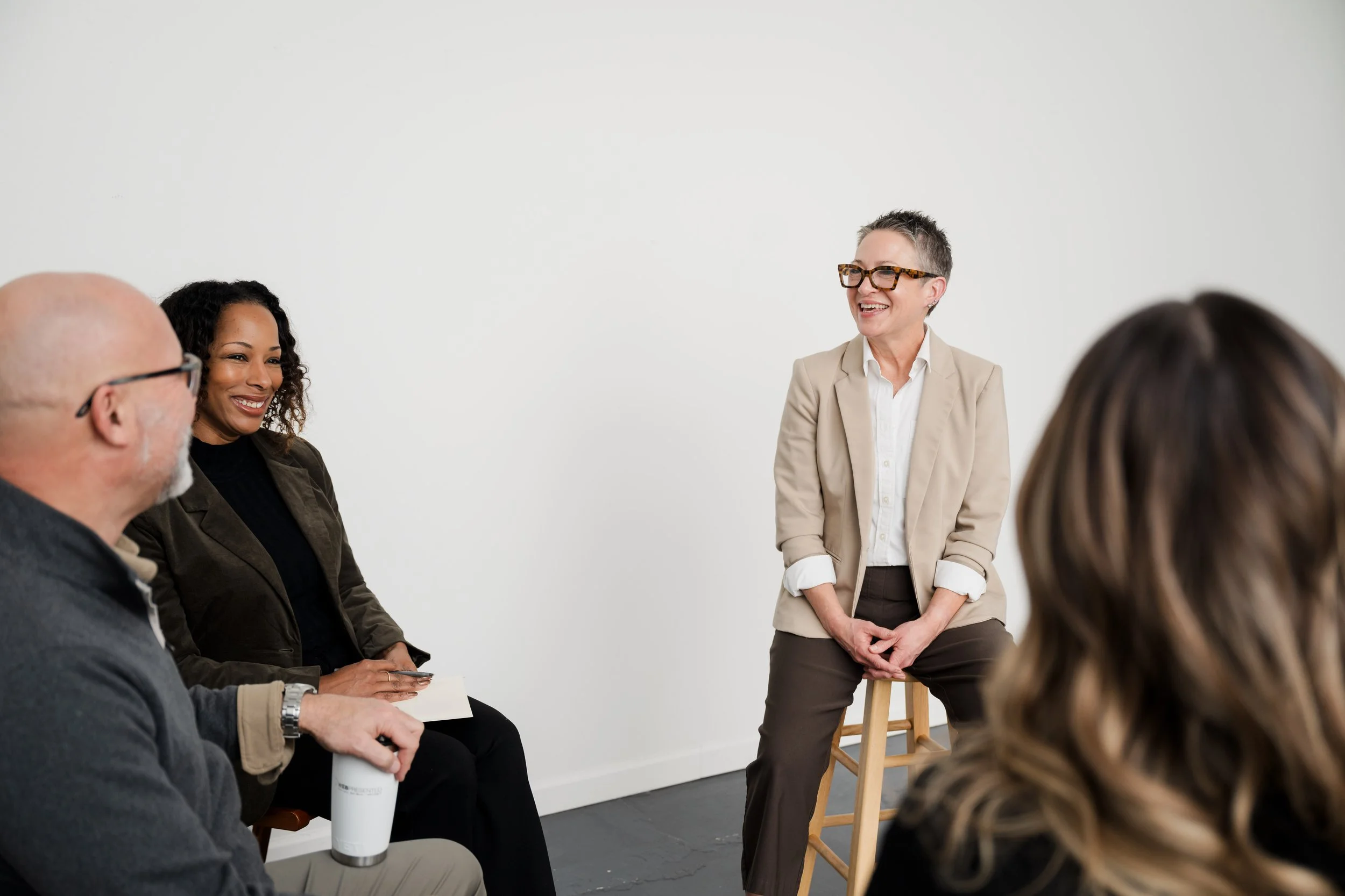 A woman in a beige blazer and glasses is standing and smiling while talking to a group of people seated in a room with a plain white wall.