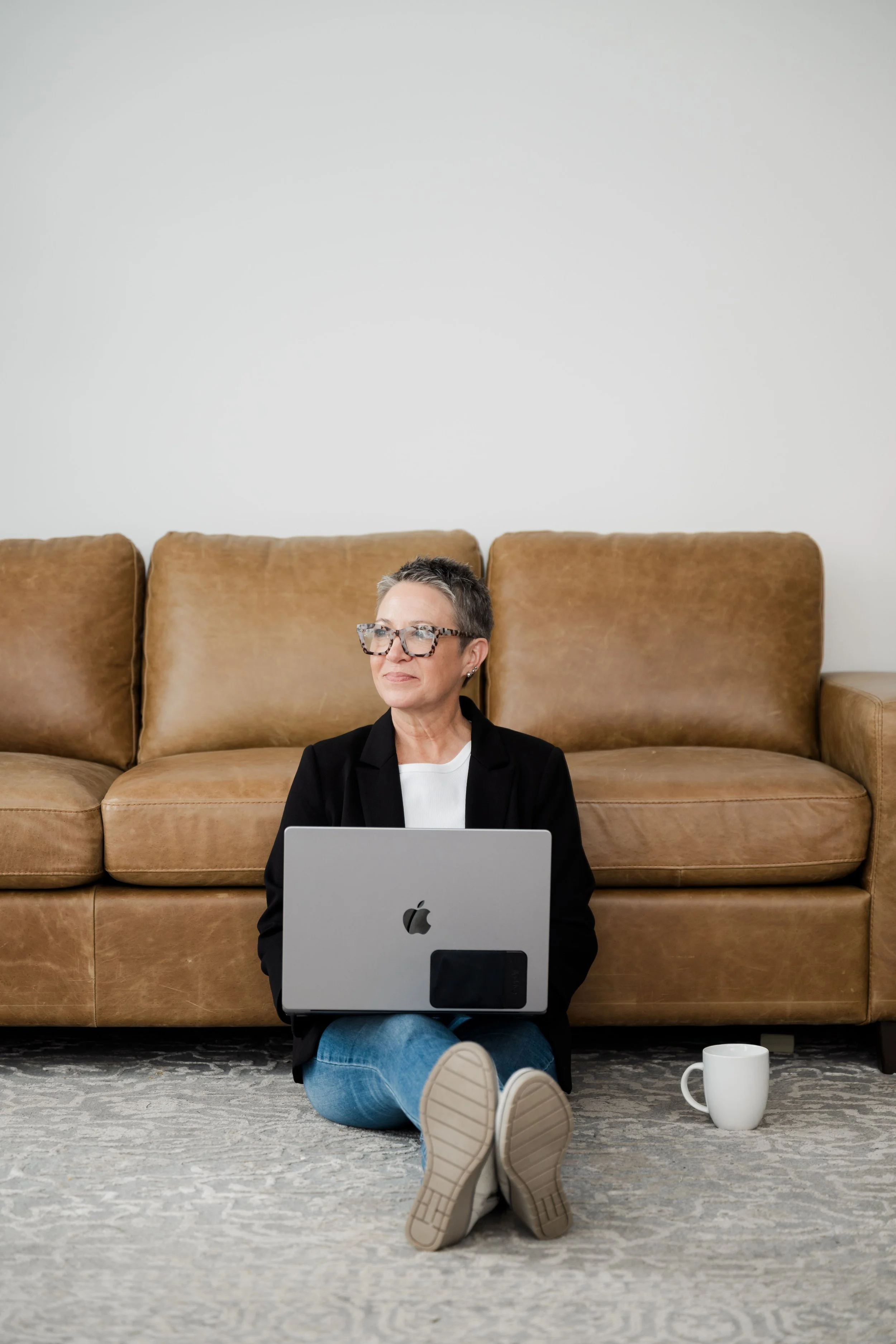 A woman with short gray hair, glasses, and a black jacket sitting on the floor with her back against a brown sofa, using a silver MacBook laptop. A white coffee mug sits next to her on the floor.