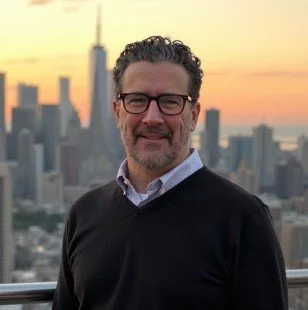 A man with glasses and a beard smiling outdoors with a city skyline and sunset in the background.