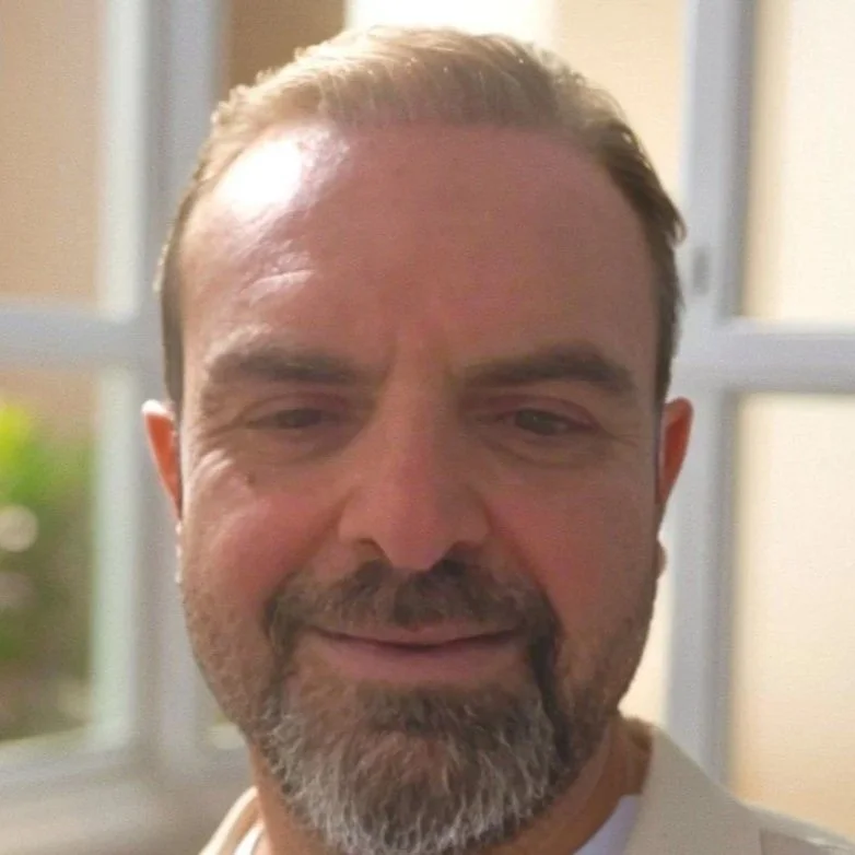 Close-up of a middle-aged man with a beard and mustache, smiling slightly, with a blurred window in the background.