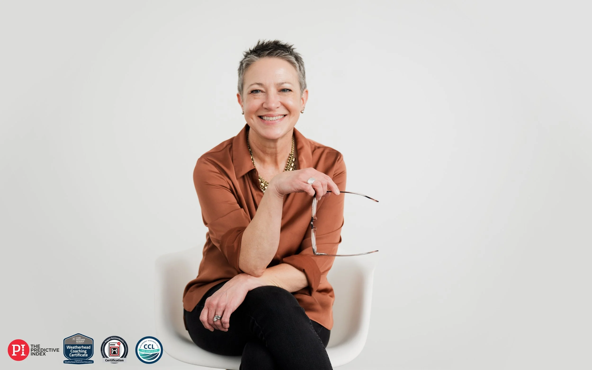 A smiling woman with short gray hair sitting on a white chair, holding eyeglasses in her right hand, wearing a brown blouse and black pants, against a plain white background.