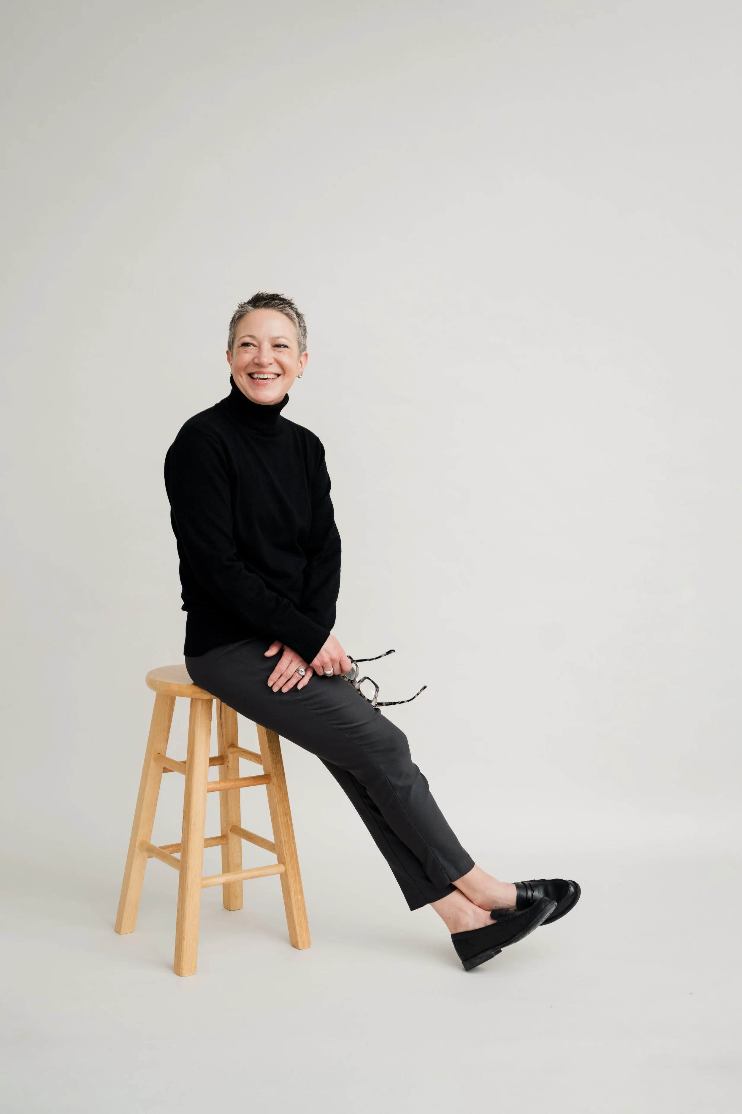 A woman with short gray hair, wearing a black turtleneck and black pants, sits on a wooden stool against a plain, light-colored background. She is smiling and holding glasses in her hand.