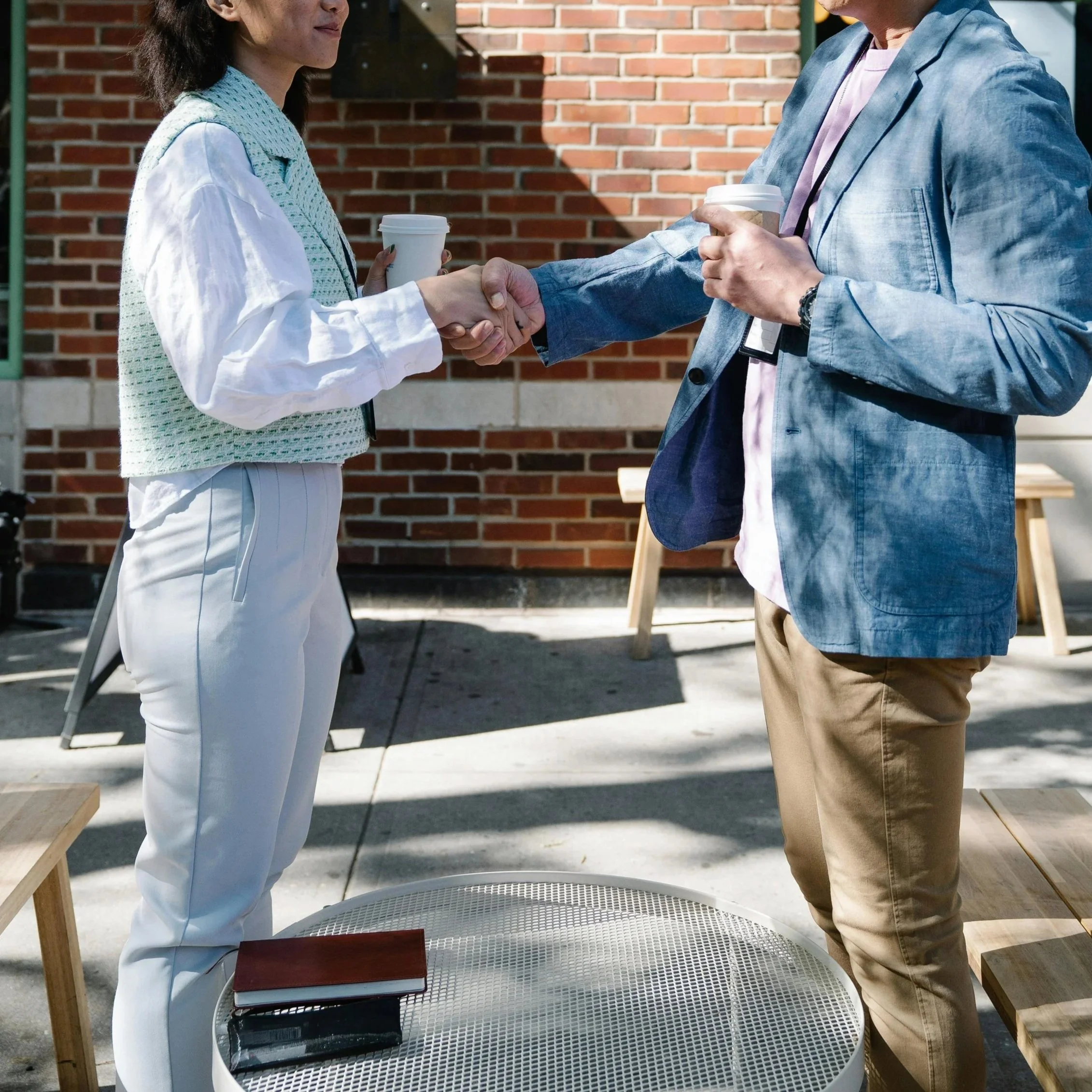 Two individuals shaking hands outdoors, each holding a disposable coffee cup, with wooden tables and a brick wall in the background.