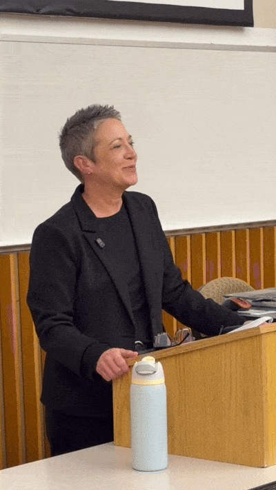 A woman with short gray hair standing at a podium during a presentation or lecture in a classroom.