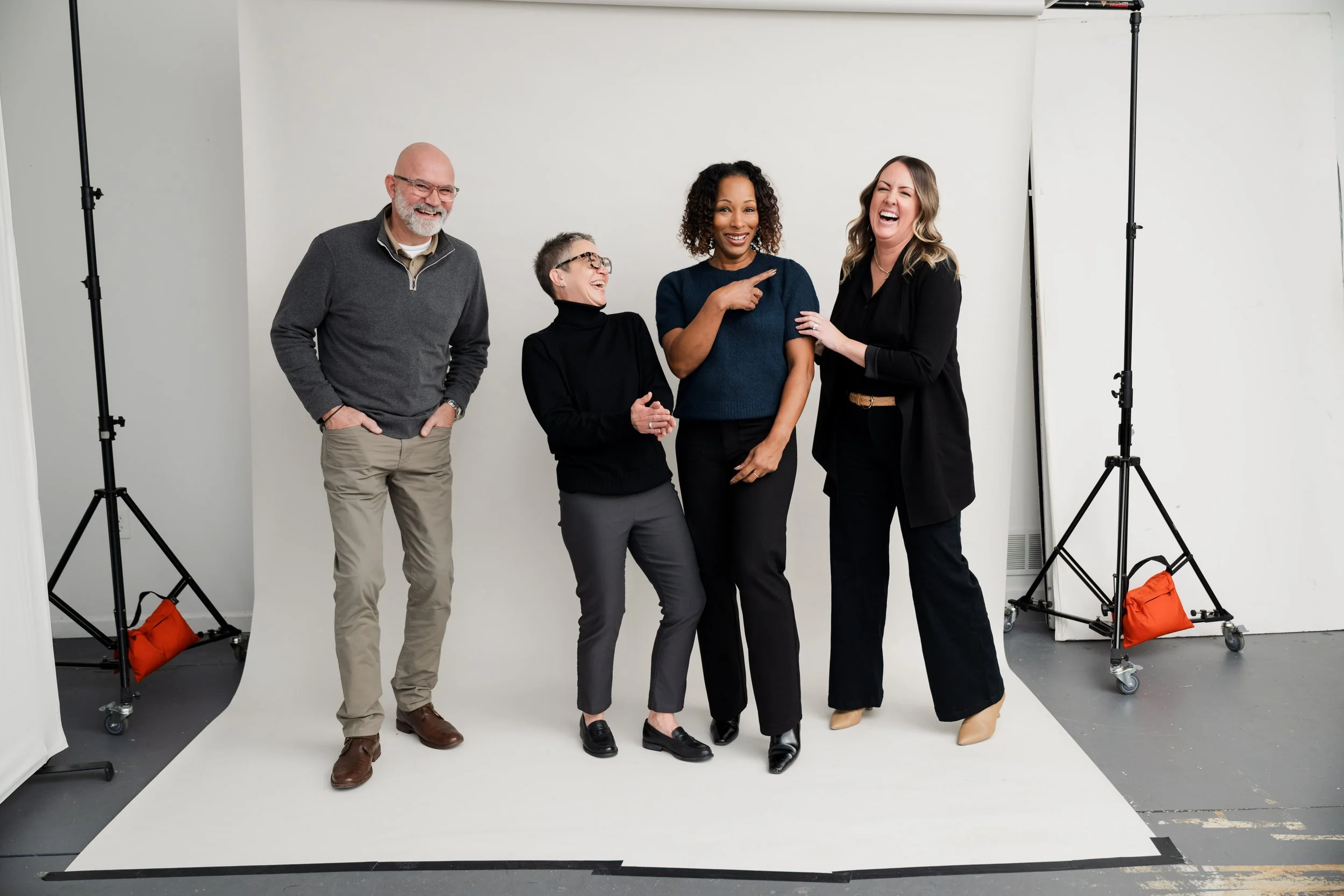 Four people standing together in a photography studio with a white backdrop, laughing and smiling. Two women and two men, dressed in business casual attire, engaging in conversation and humor.