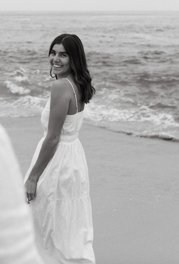 A woman in a white dress smiling and standing on a beach, with the ocean in the background.