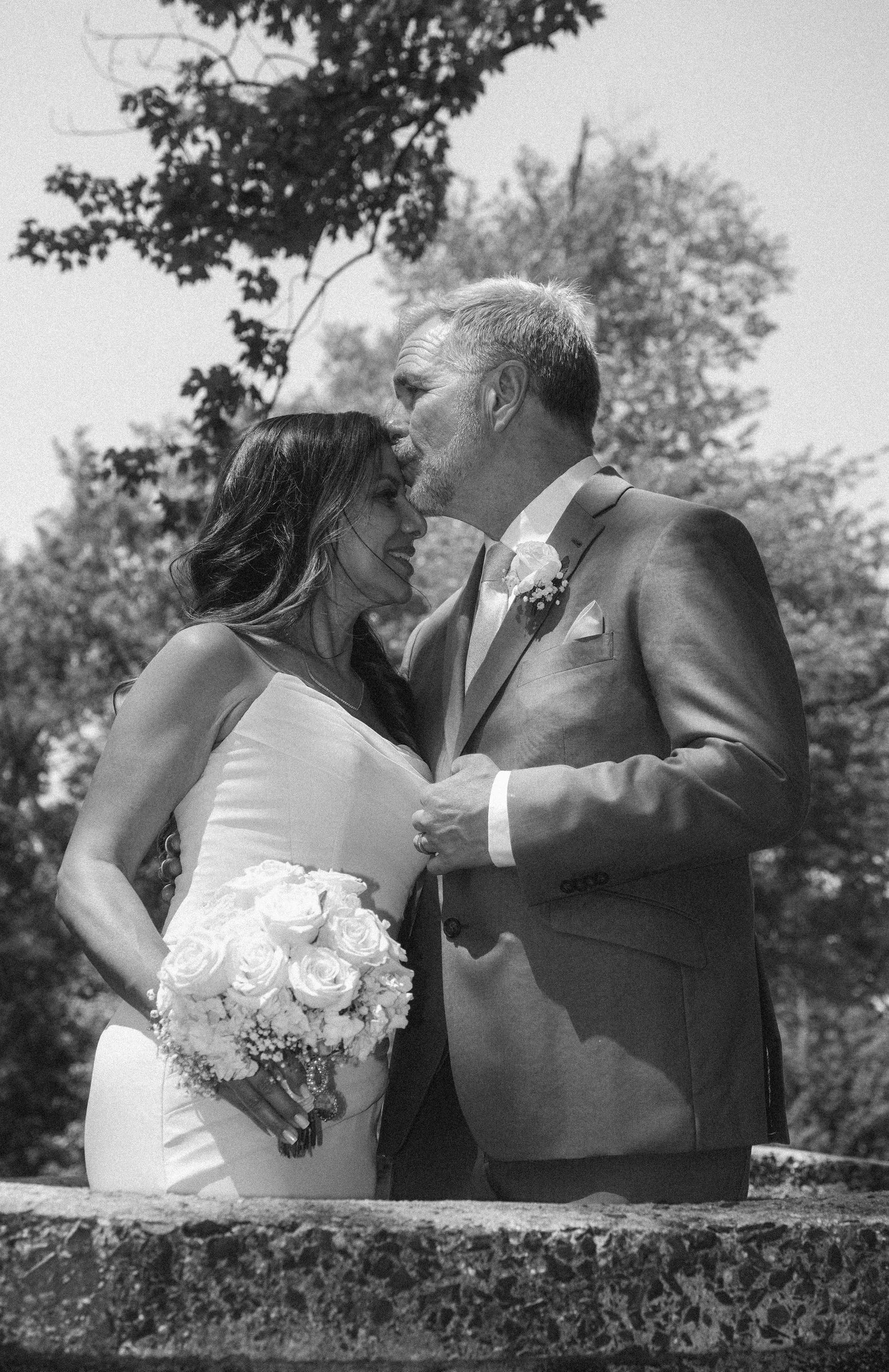 A black and white photo of a bride and groom on their wedding day. The groom is kissing the bride's forehead while holding her hands. The bride is holding a bouquet of white roses and is smiling. The scene is outdoors with trees in the background.