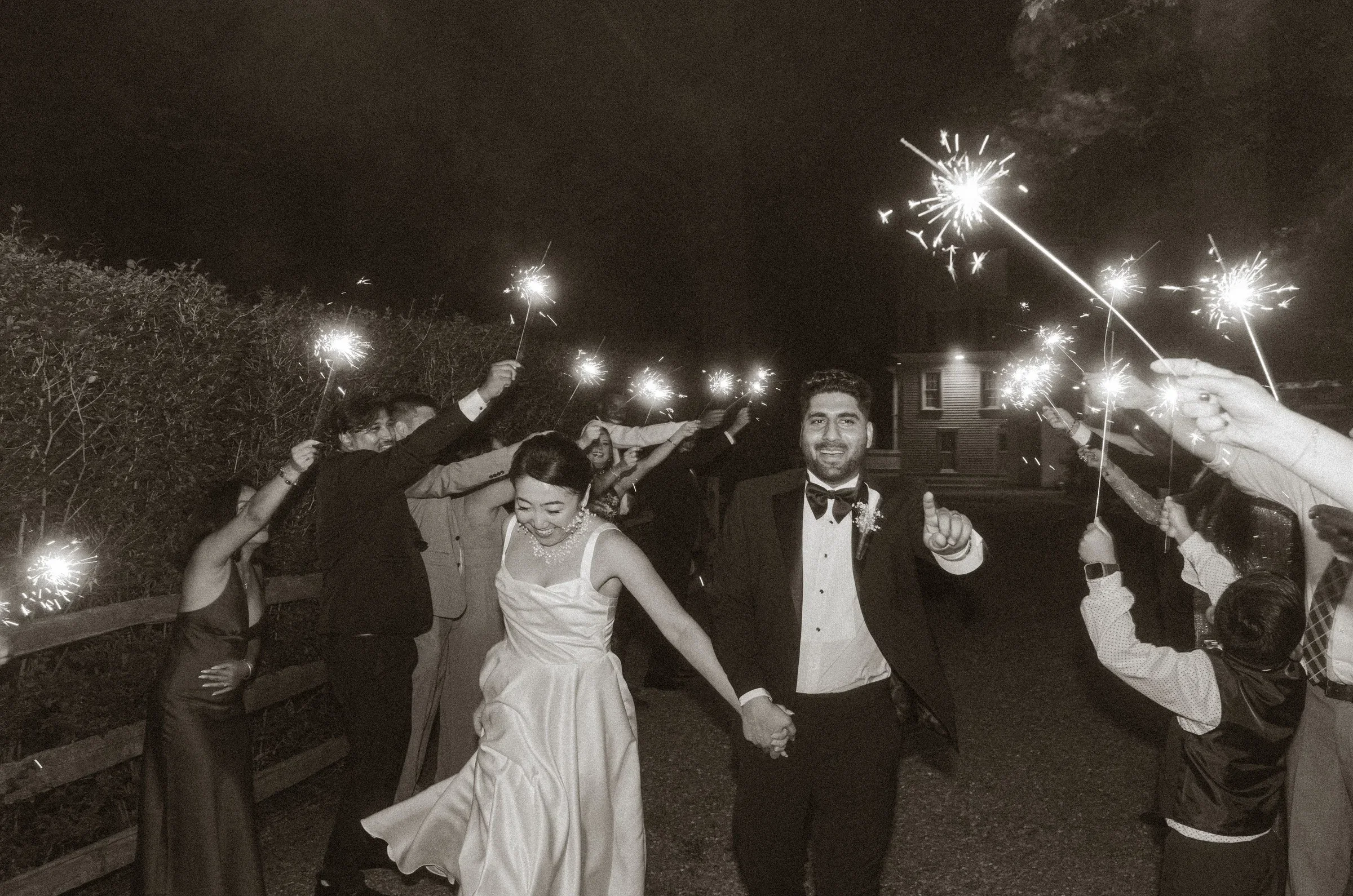 People celebrating at night with sparklers, including a man and woman in formal attire, holding hands and smiling, outdoors near a house and trees.