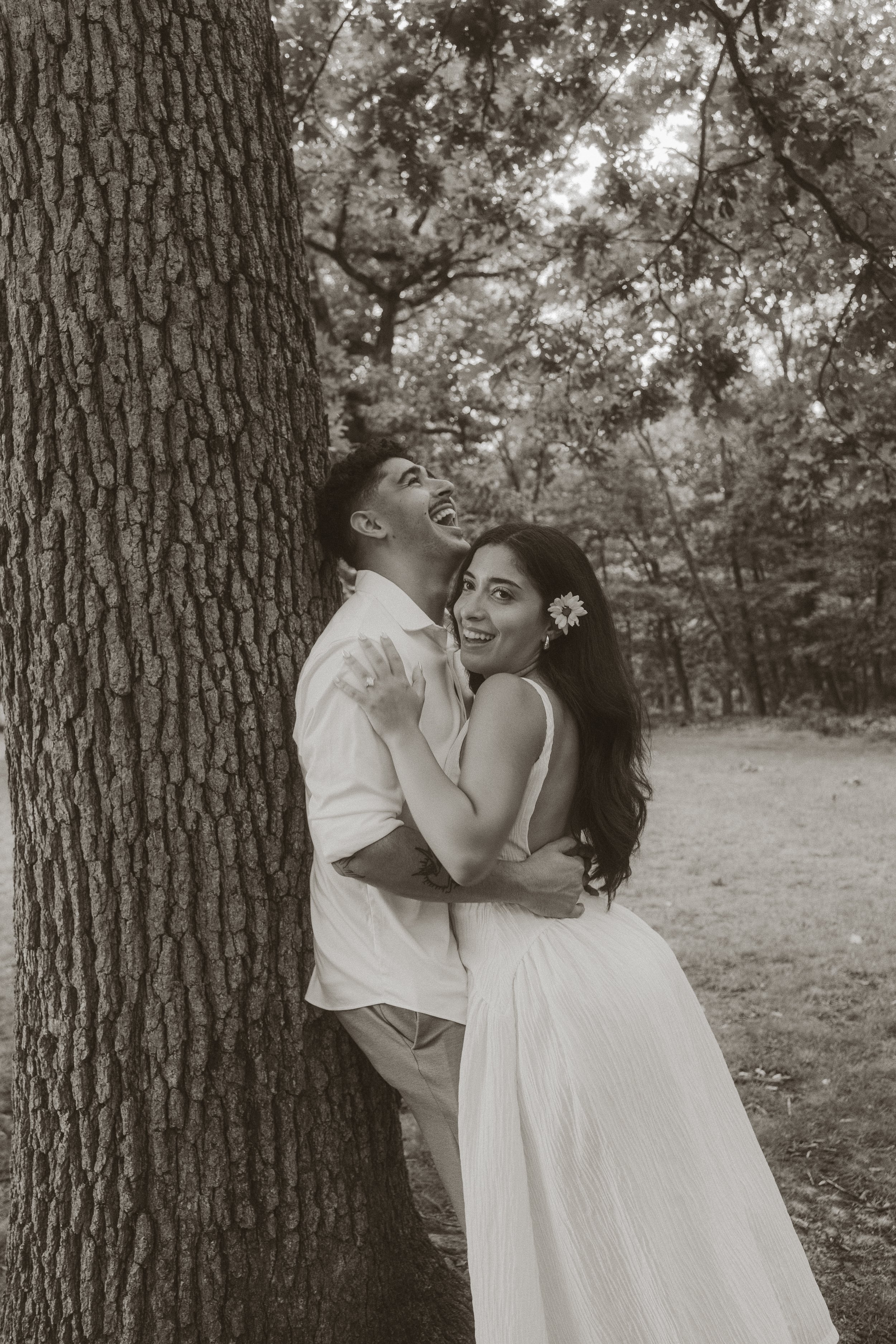 A joyful young couple in formal attire embracing outdoors, with a large tree trunk in the foreground and trees in the background.