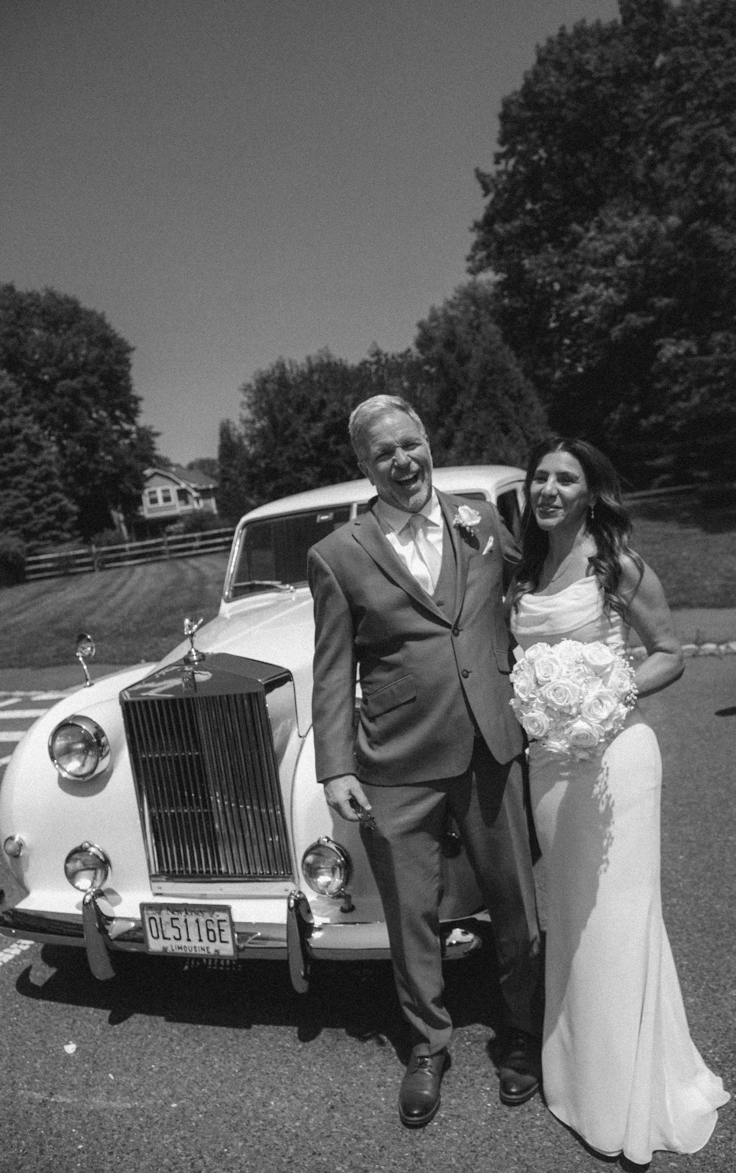 A happy newlywed couple standing in front of a vintage white car outdoors on a sunny day. The groom is wearing a suit and the bride is in a wedding dress holding a bouquet of roses. Trees and a house are in the background.