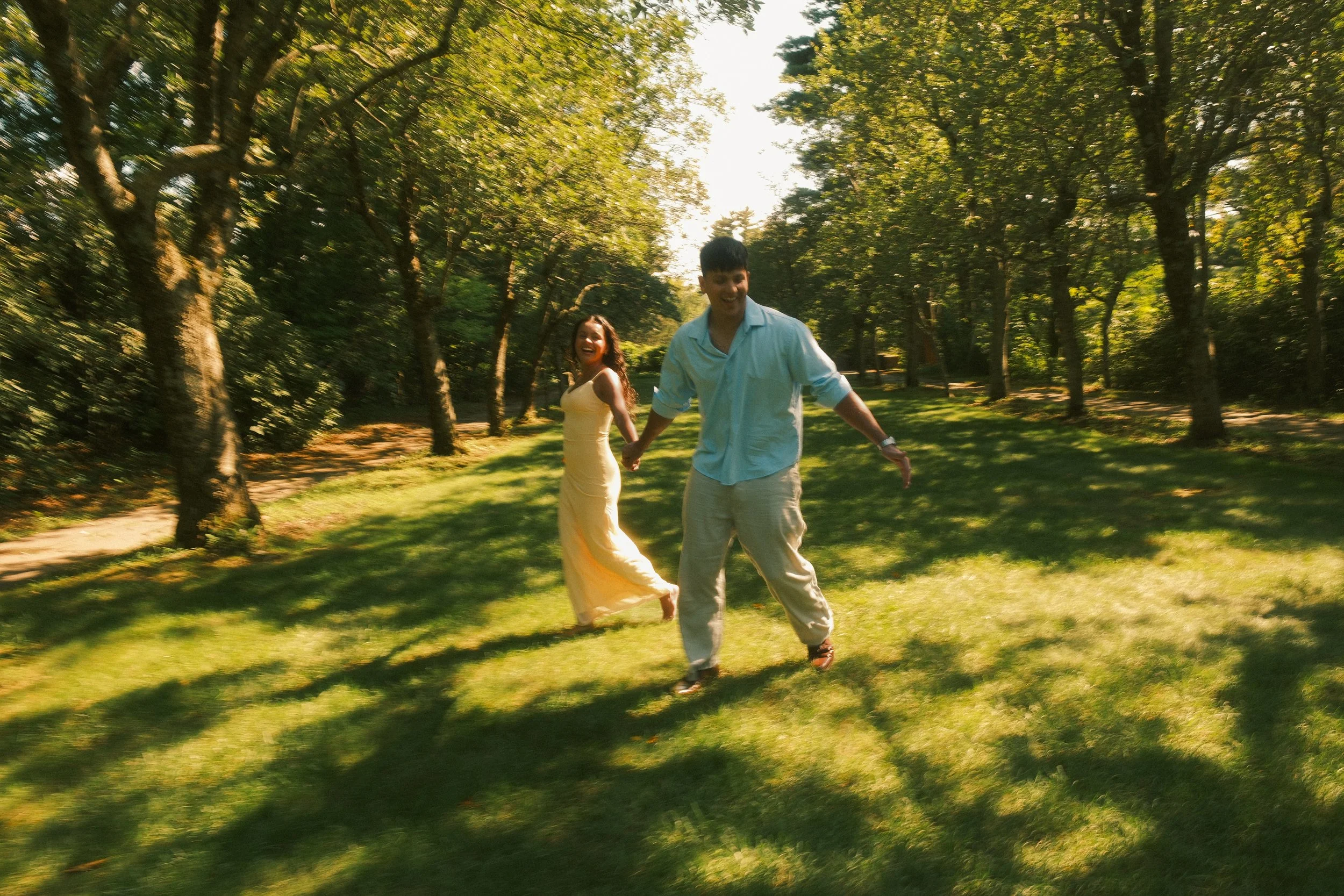 A smiling couple holding hands and walking through a sunlit park with green grass and tall trees.
