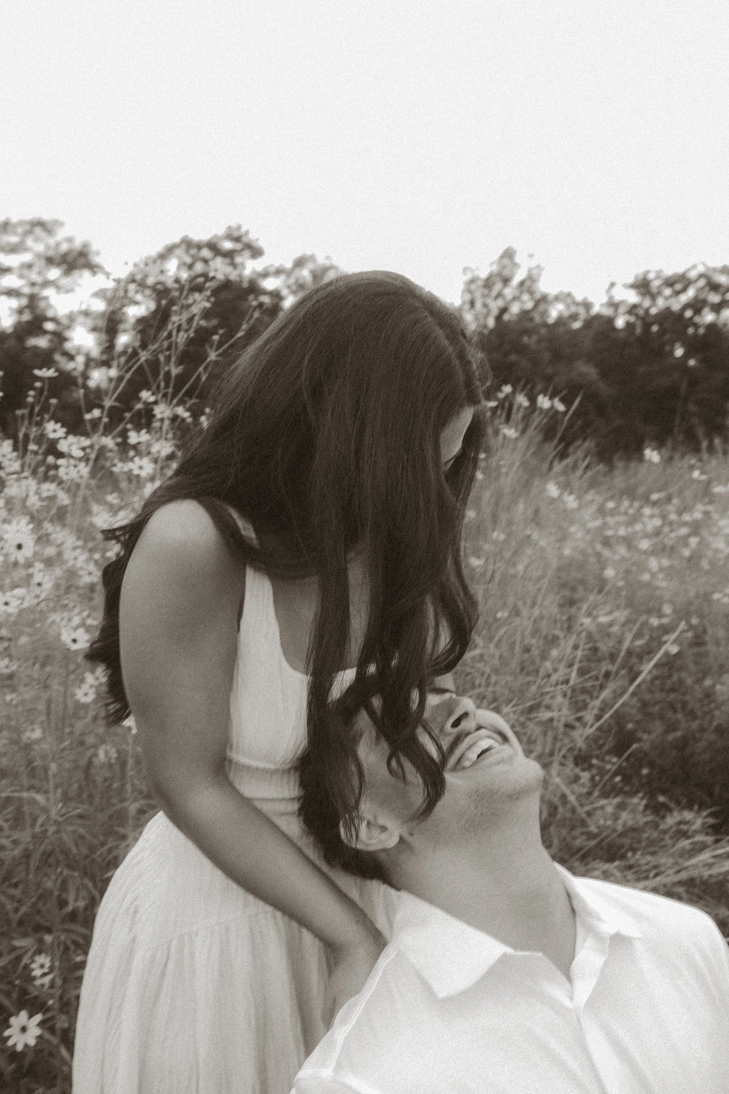 A woman with long dark hair in a white dress is sitting in a field of wildflowers, with a man lying on her lap, smiling up at her. Trees are visible in the background, and the image is in black and white.