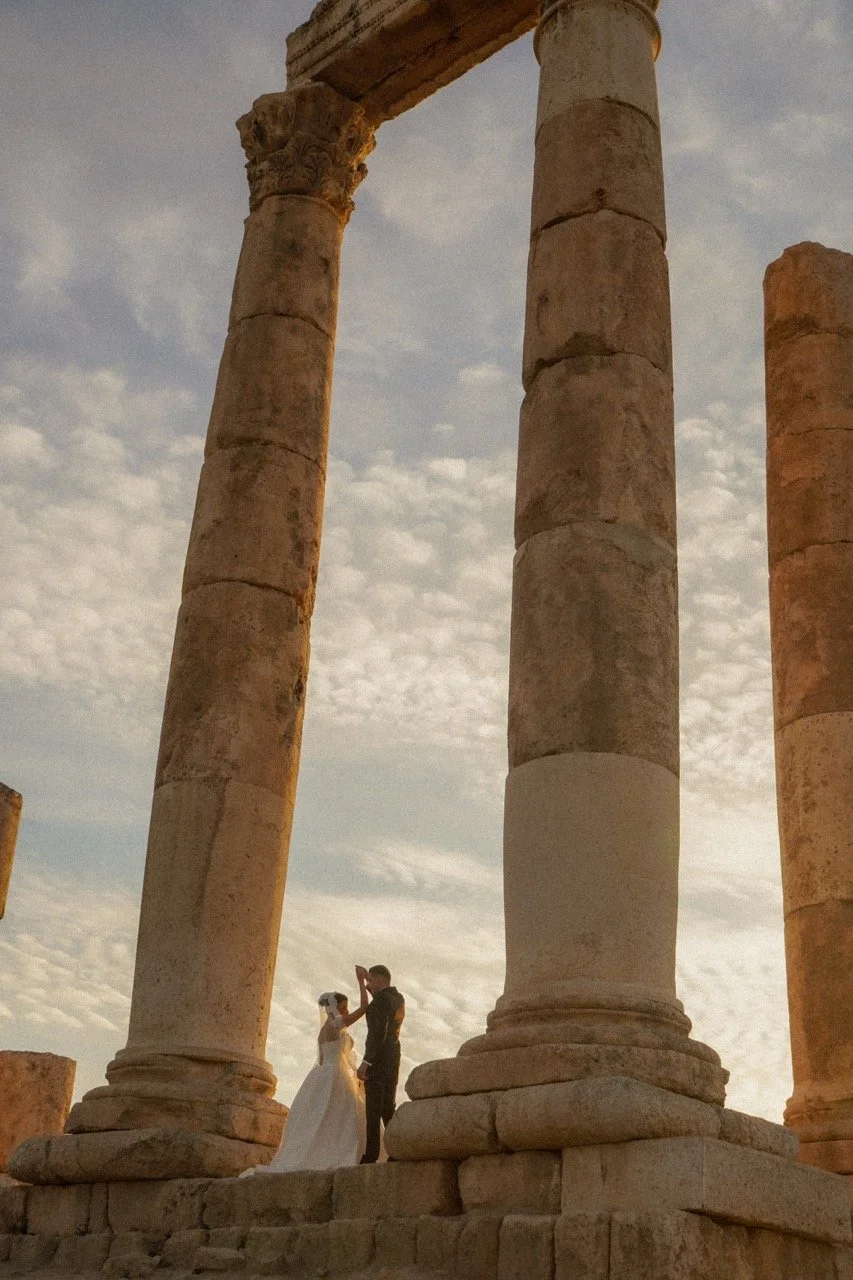 A bride and groom standing between ancient stone columns during sunset.