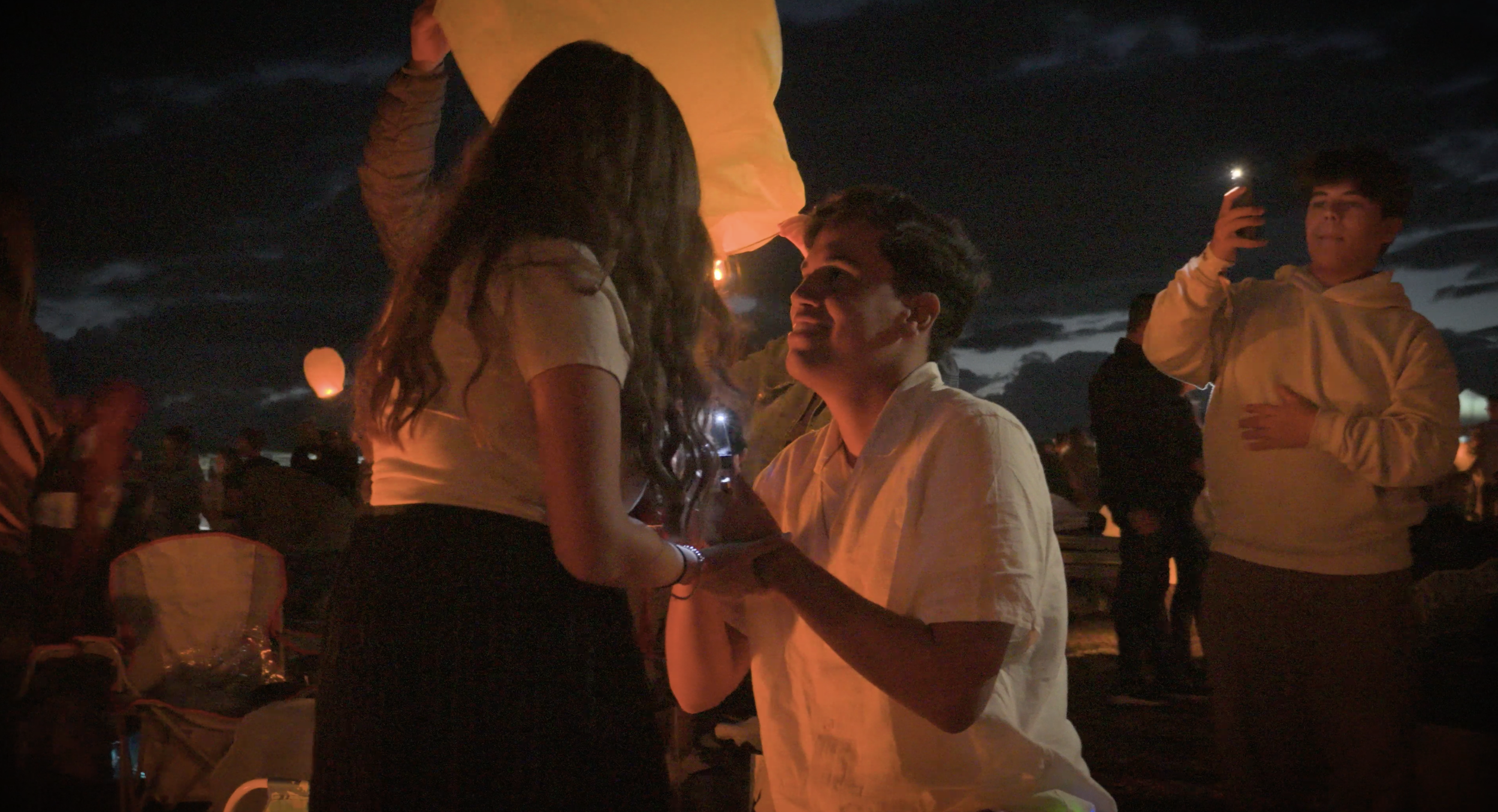 A young man is proposing marriage to a young woman during a night outdoor event, with other people in the background taking photos and watching.
