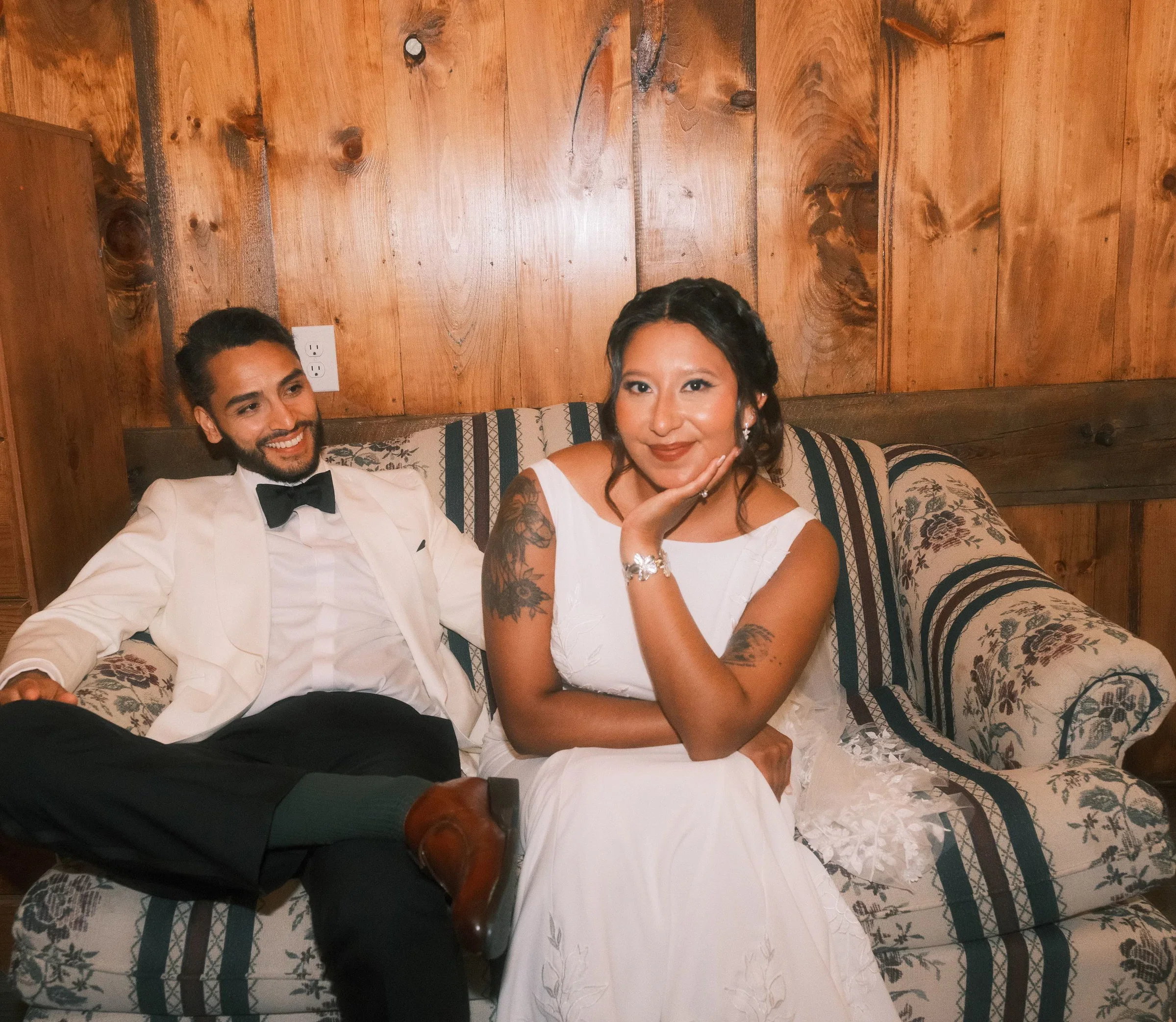 A man in formal attire, a woman in a white dress, sitting on a floral-patterned couch against a wooden-paneled wall, smiling and posing for a photo.