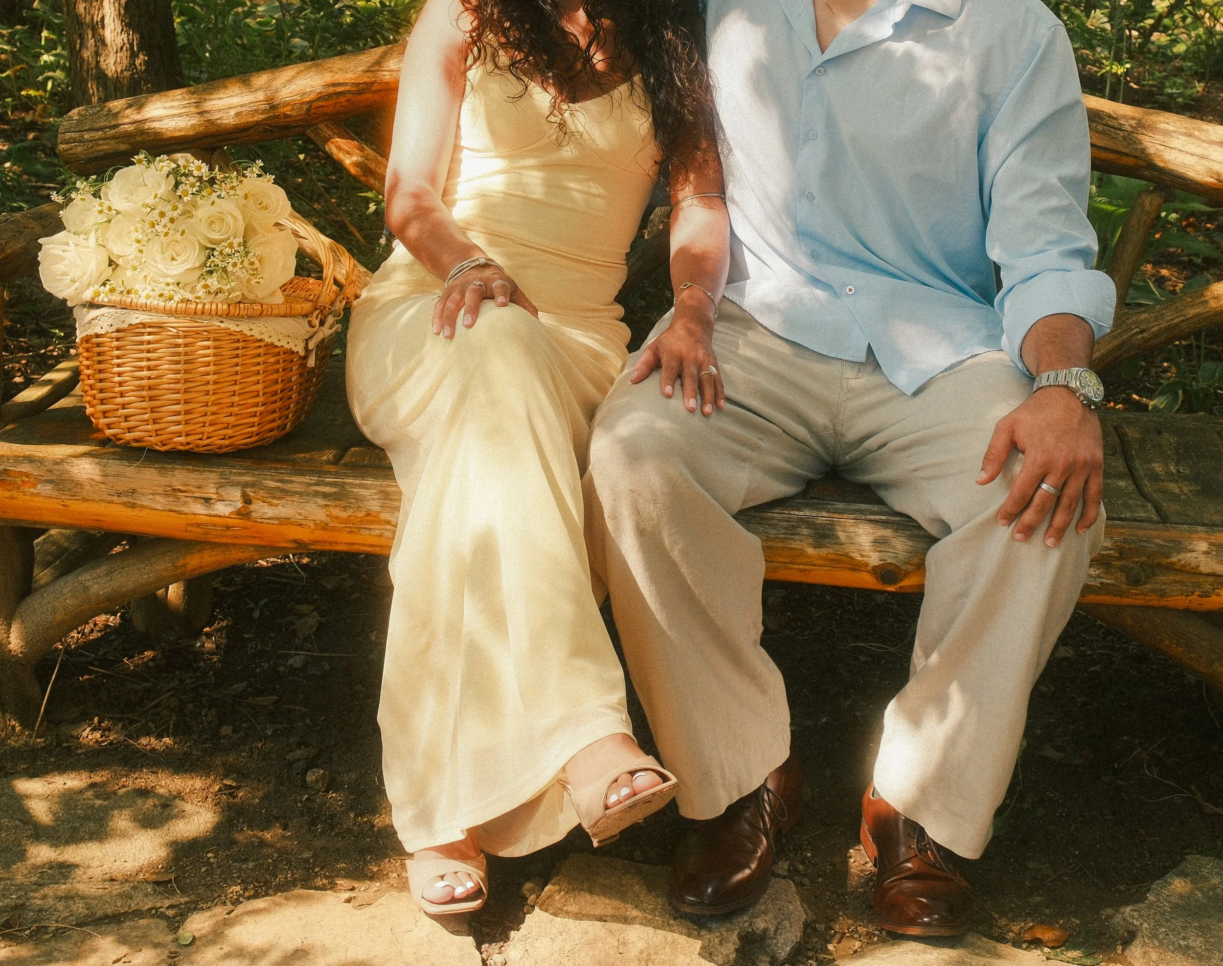 A couple sitting together on a wooden bench outdoors. The woman is wearing a light yellow dress, and the man is wearing a light blue shirt and beige pants. There is a wicker basket with white roses and daisies beside them, and both are displaying wed