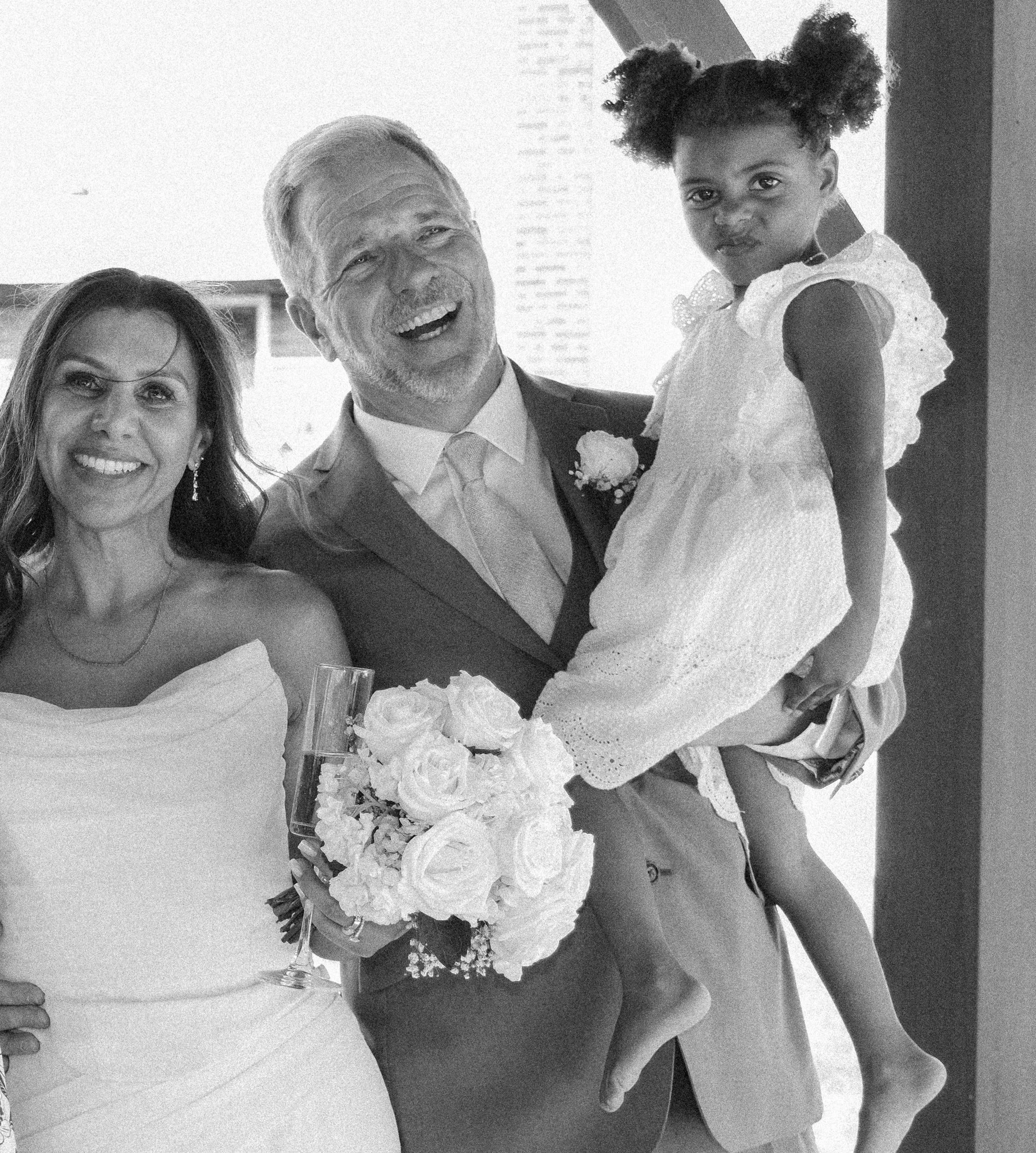 A black and white photo of a man in a suit and tie holding a young girl in a dress, smiling at a celebration or formal event. A woman with long dark hair is partially visible on the left side of the image. The man appears happy and is standing next t