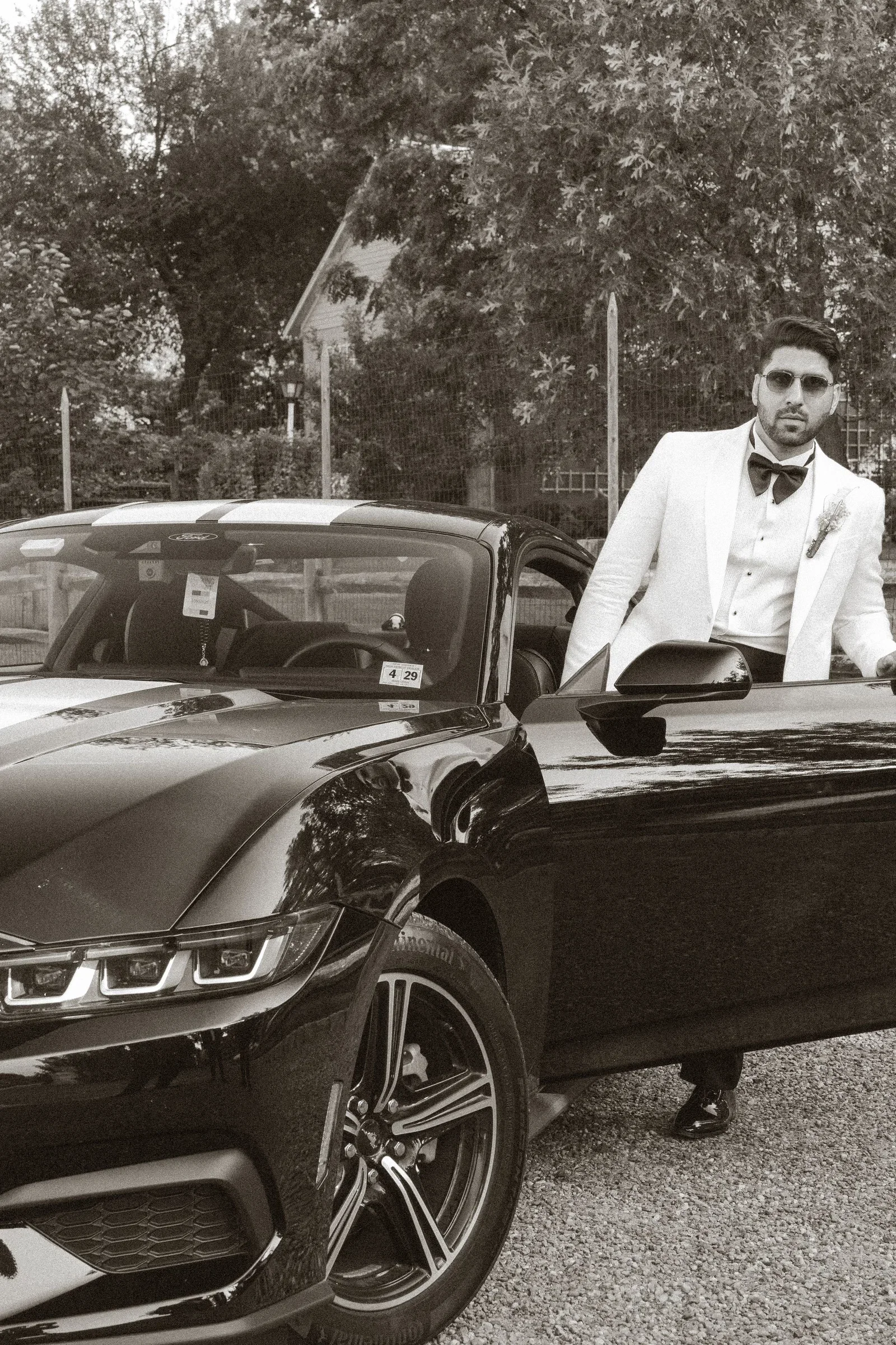 A man in formal attire with sunglasses, a white tuxedo, black bow tie, and boutonnière, standing next to a black sports car with white racing stripes, outdoors with trees and a fence in the background.