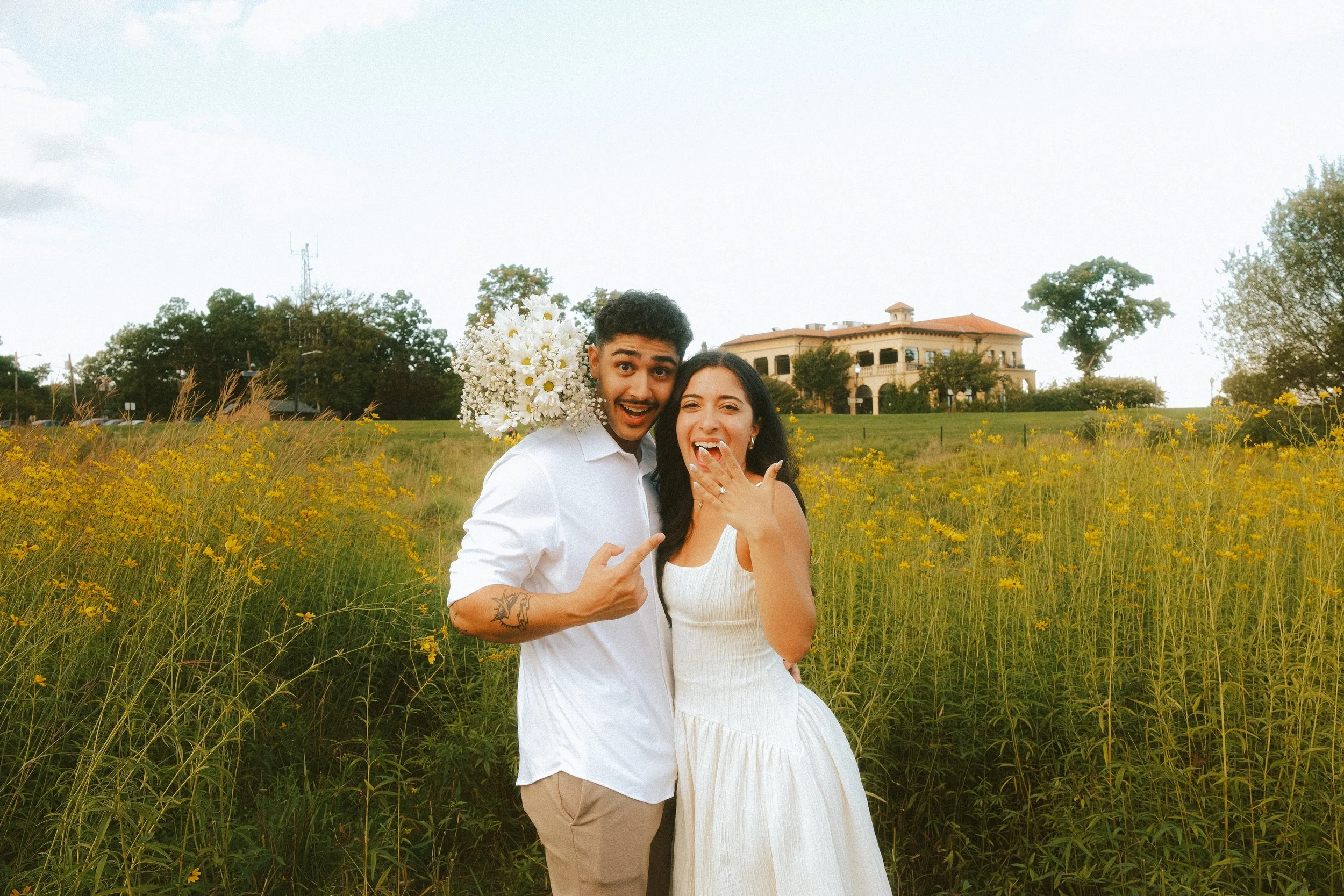A happy couple standing in a field of yellow flowers with a large, elegant house in the background. The man is holding a bouquet of white daisies and wearing a white shirt, while the woman is wearing a white dress. They appear joyful and celebratory.