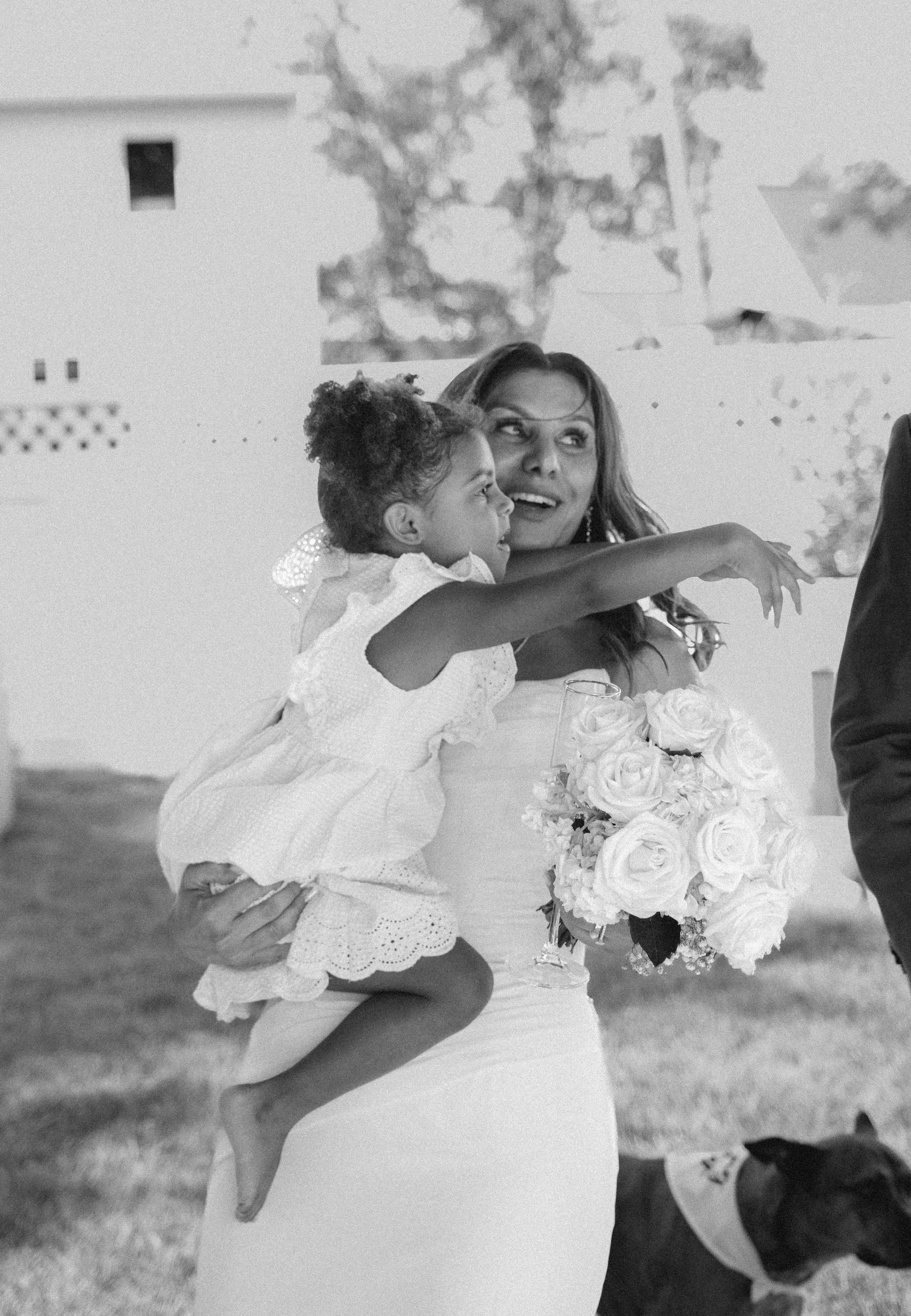 Woman holding a young girl with curly hair, both dressed in white, at an outdoor event with a large bouquet of white roses in front of them.