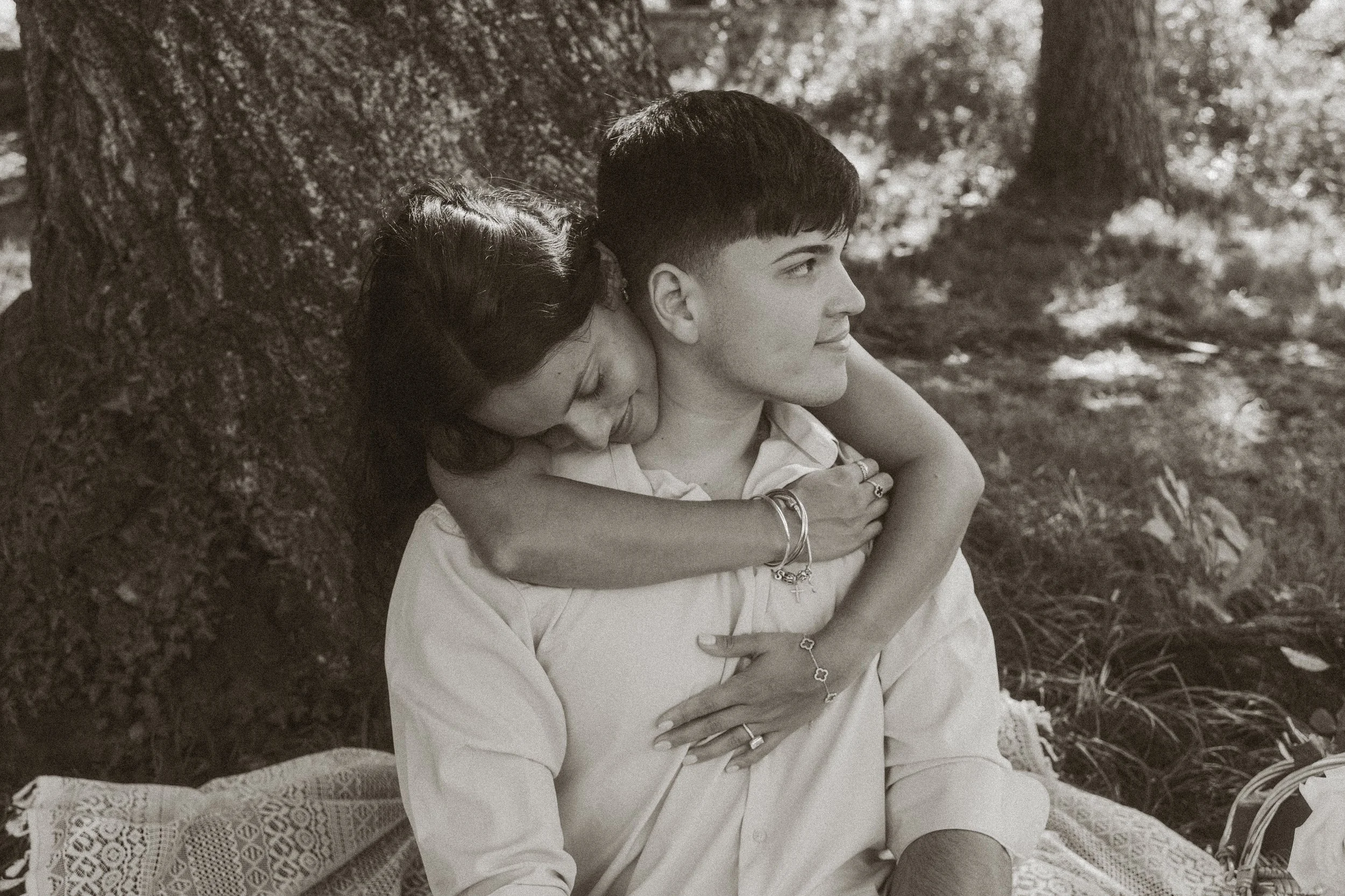 A young man and woman sitting outdoors by a tree, the woman resting her head on the man's shoulder, with their arms intertwined. The scene appears peaceful and intimate.