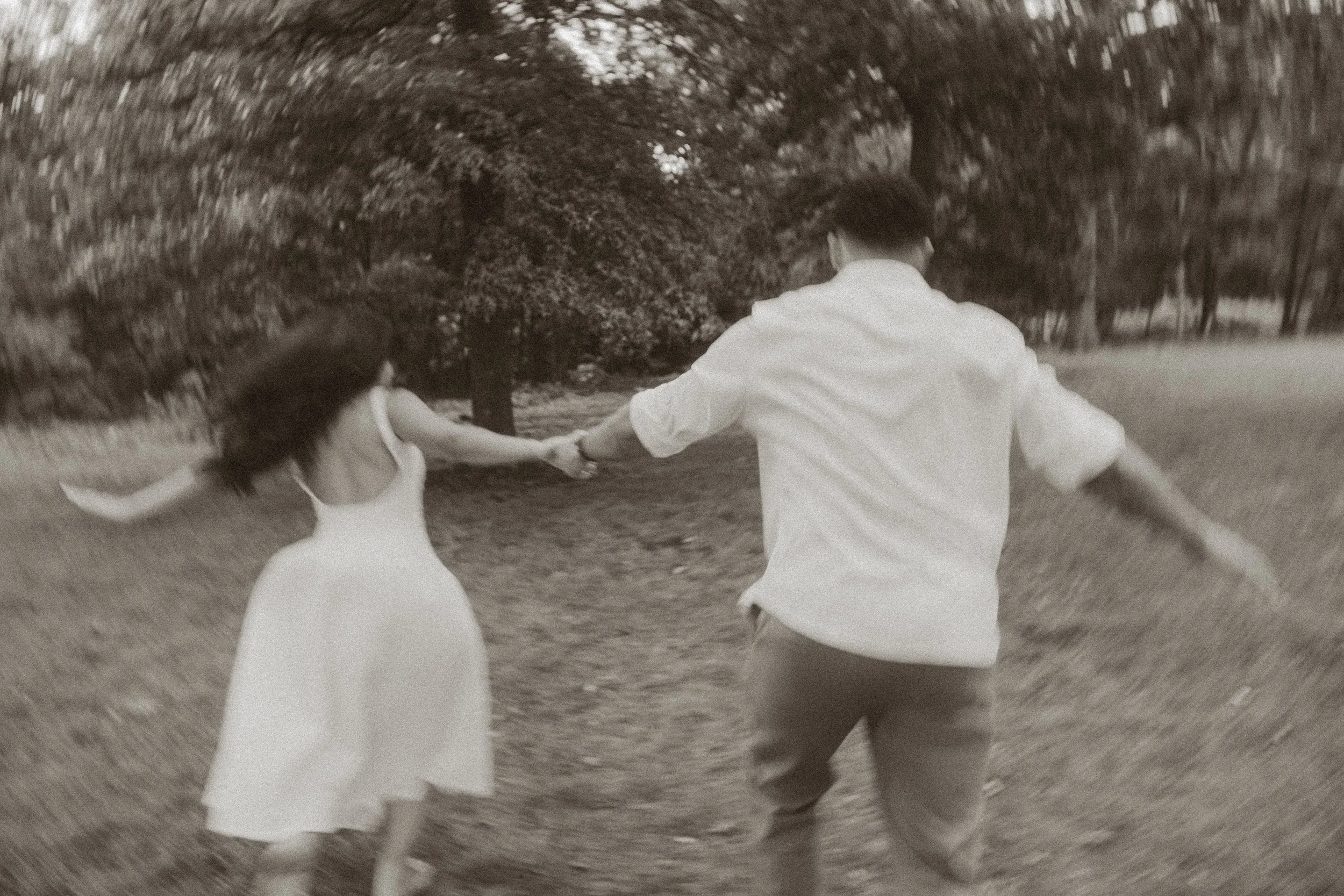 A man and a woman are holding hands and spinning around outdoors in a park with trees in the background.
