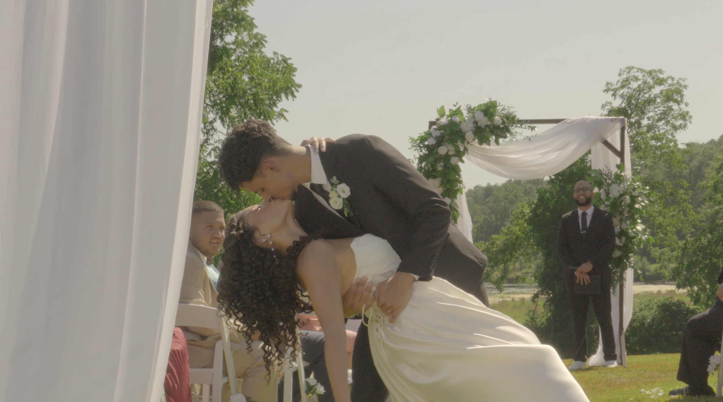 A couple shares a kiss during their outdoor wedding ceremony, with a floral arch and officiant visible in the background. The groom is dipping the bride as they kiss, surrounded by friends and greenery.