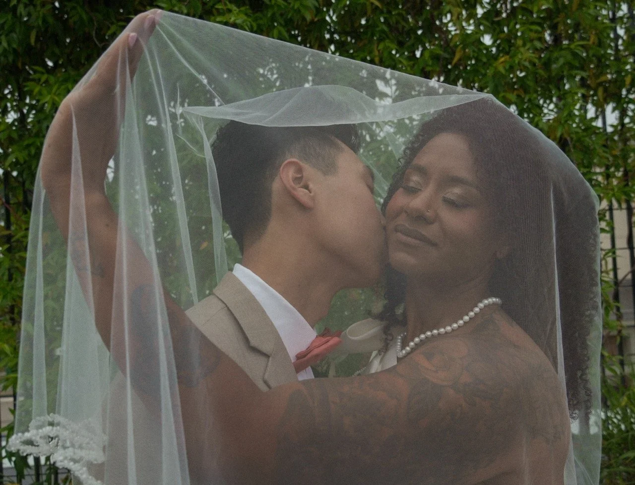 A bride and groom sharing a kiss under a wedding veil outdoors, surrounded by green foliage.